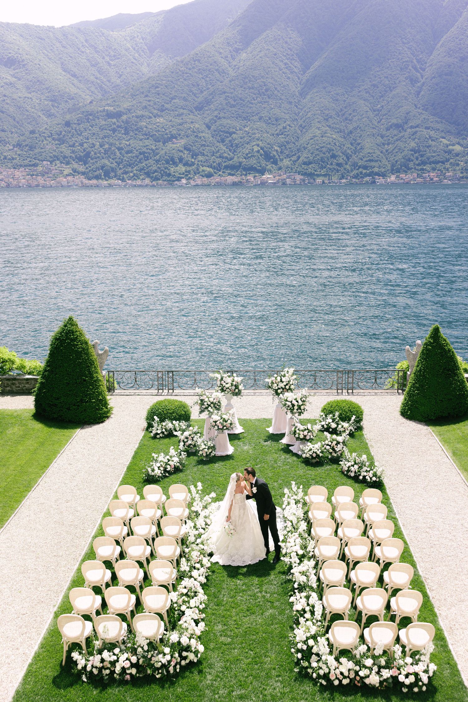 Lakeside wedding ceremony setup with white chairs arranged in curved rows and manicured topiary against mountain views.
