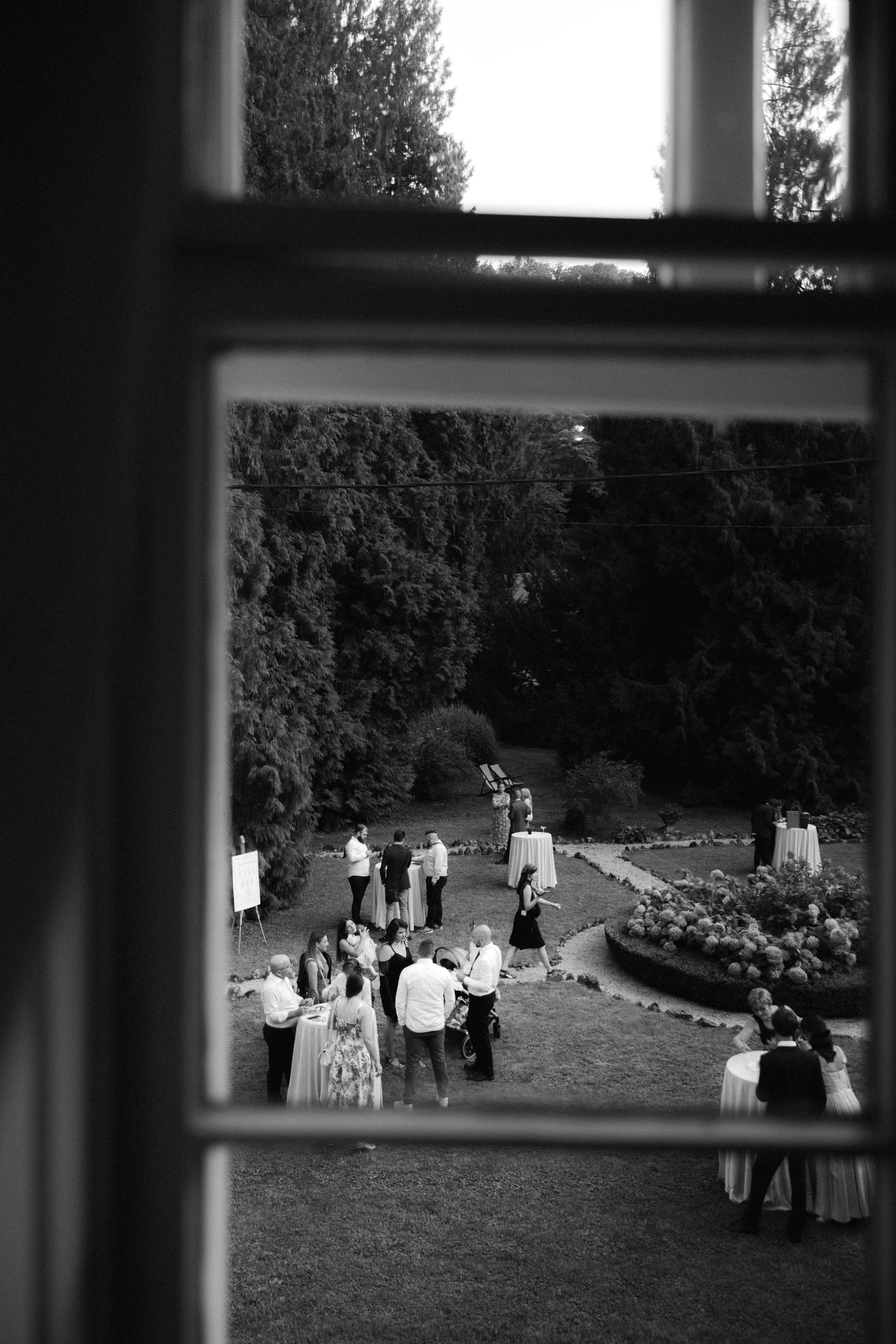 Black and white view through a window of wedding guests mingling on a garden lawn during an outdoor reception.