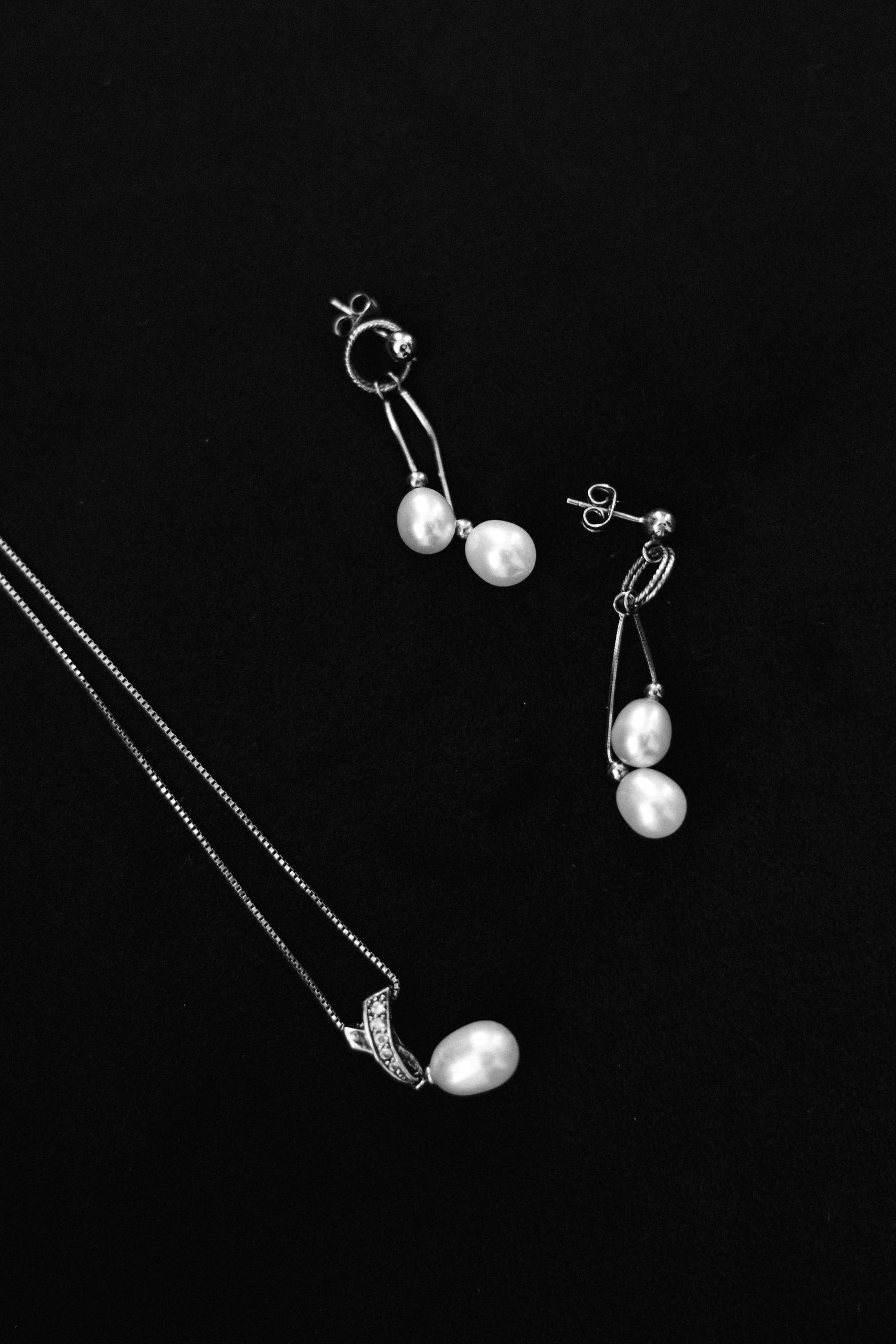 A collection of delicate silver jewelry pieces including pearl earrings and necklaces displayed on a black background.