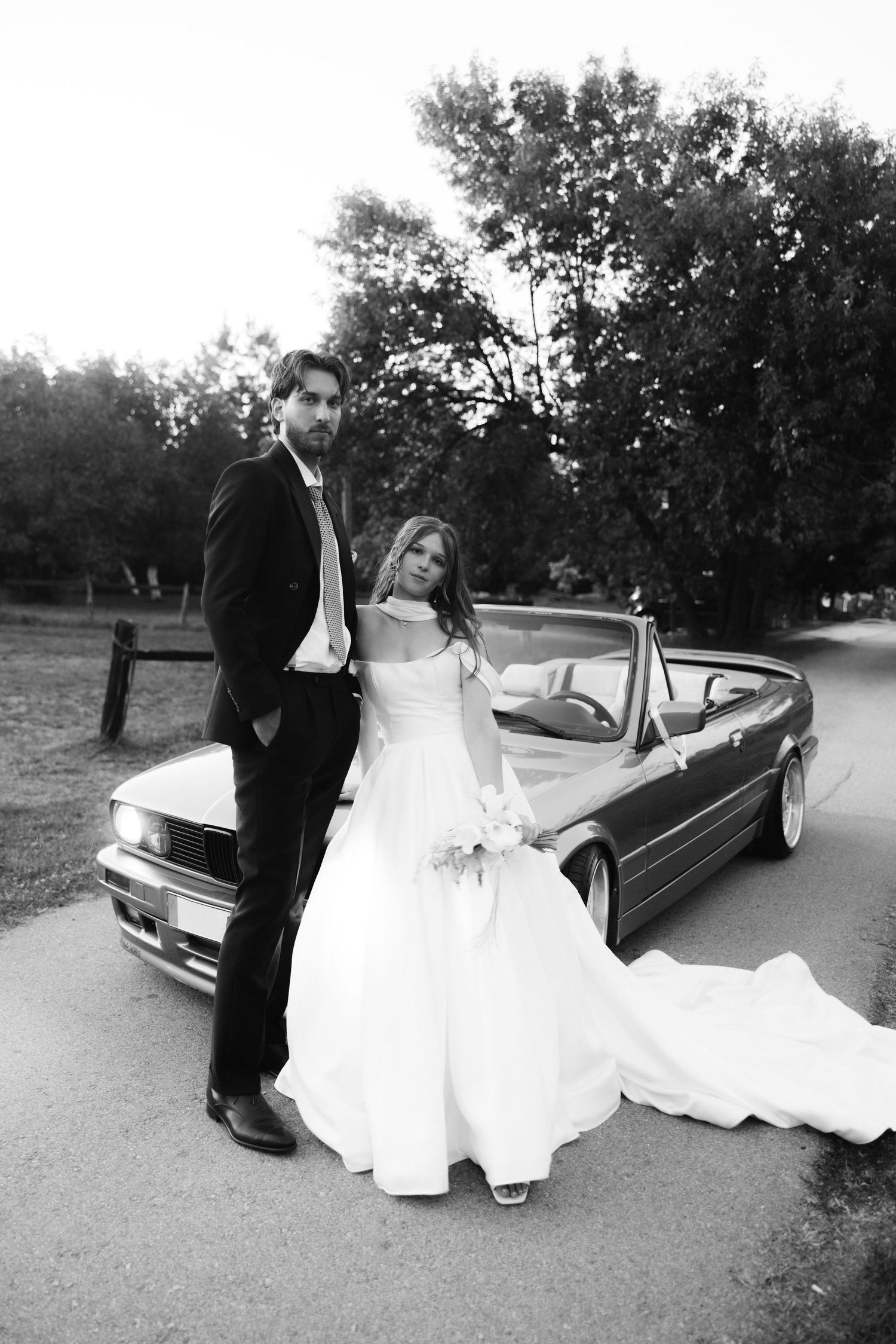 A black and white wedding photo series showing a couple posing with a classic car in an outdoor setting.