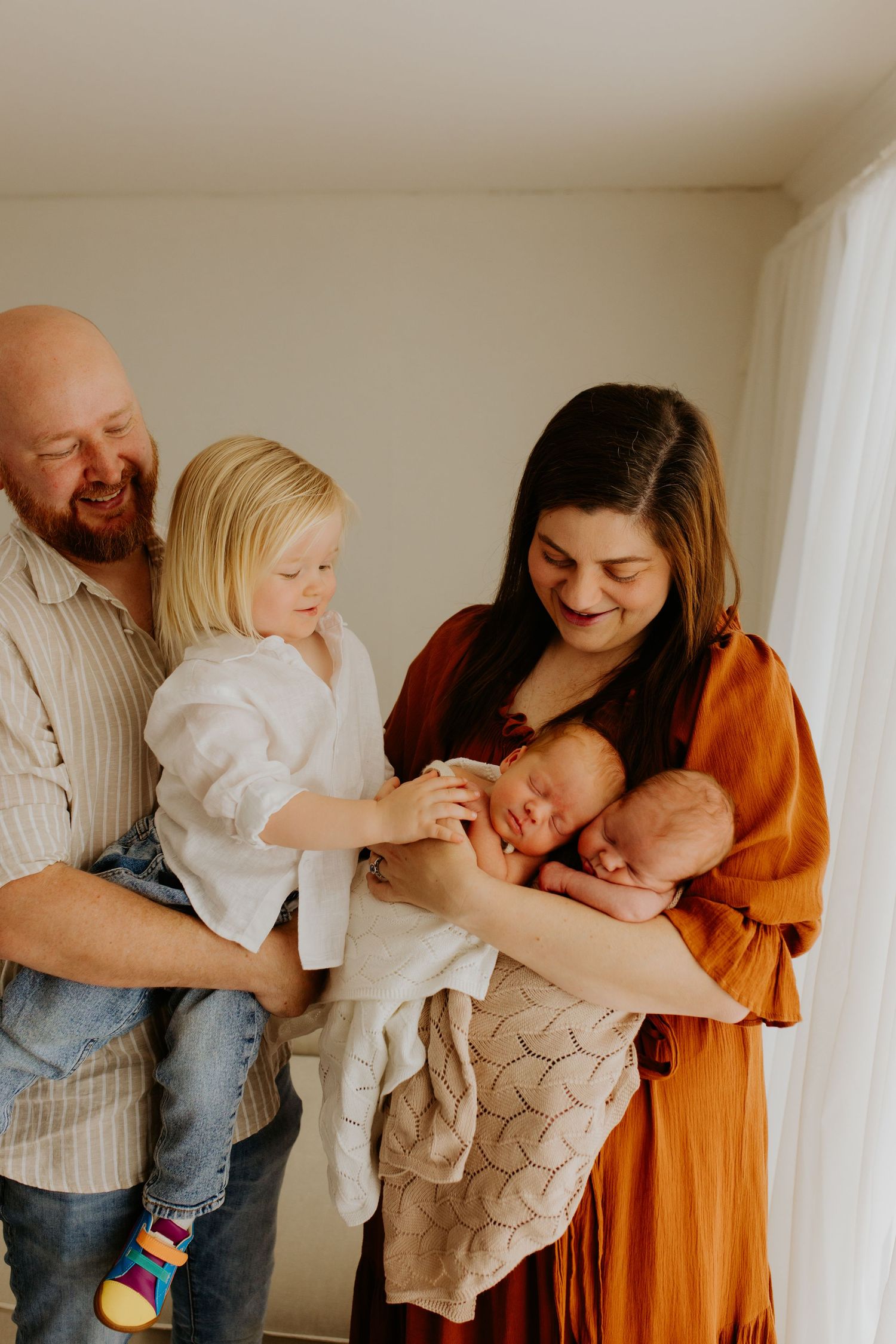 A joyful family embraces their newborn twins, with their toddler sibling reaching out with curiosity and love, showcasin