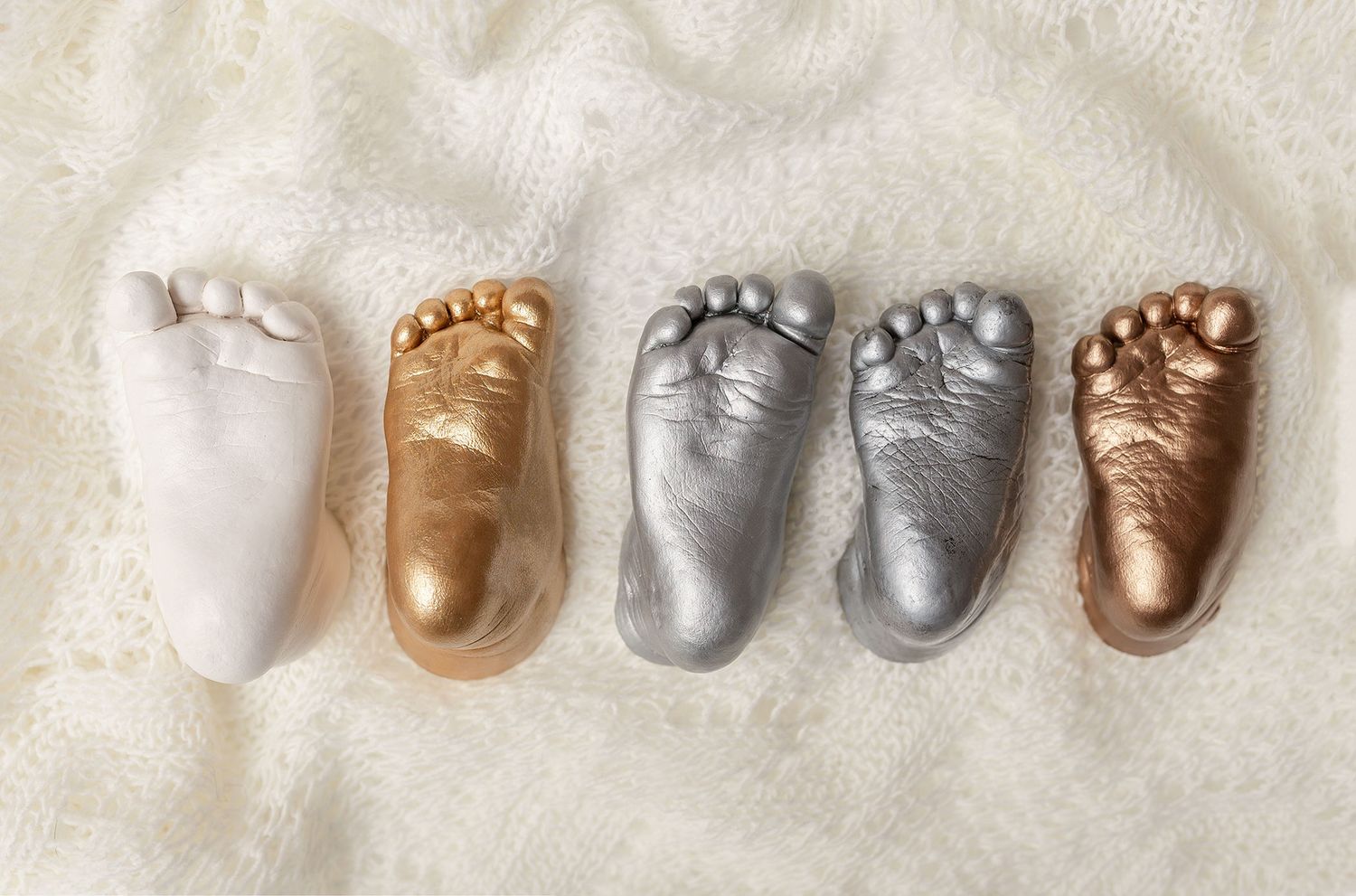 A collection of metallic gold, silver, and bronze baby foot castings arranged in a row on white fabric.