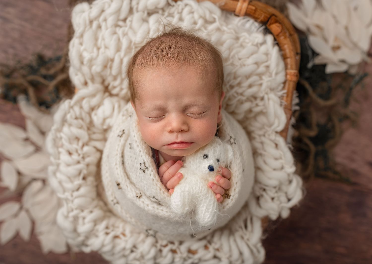 Sleeping newborn wrapped in white knit swaddle lying in cozy cream-colored basket with textured blanket underneath.