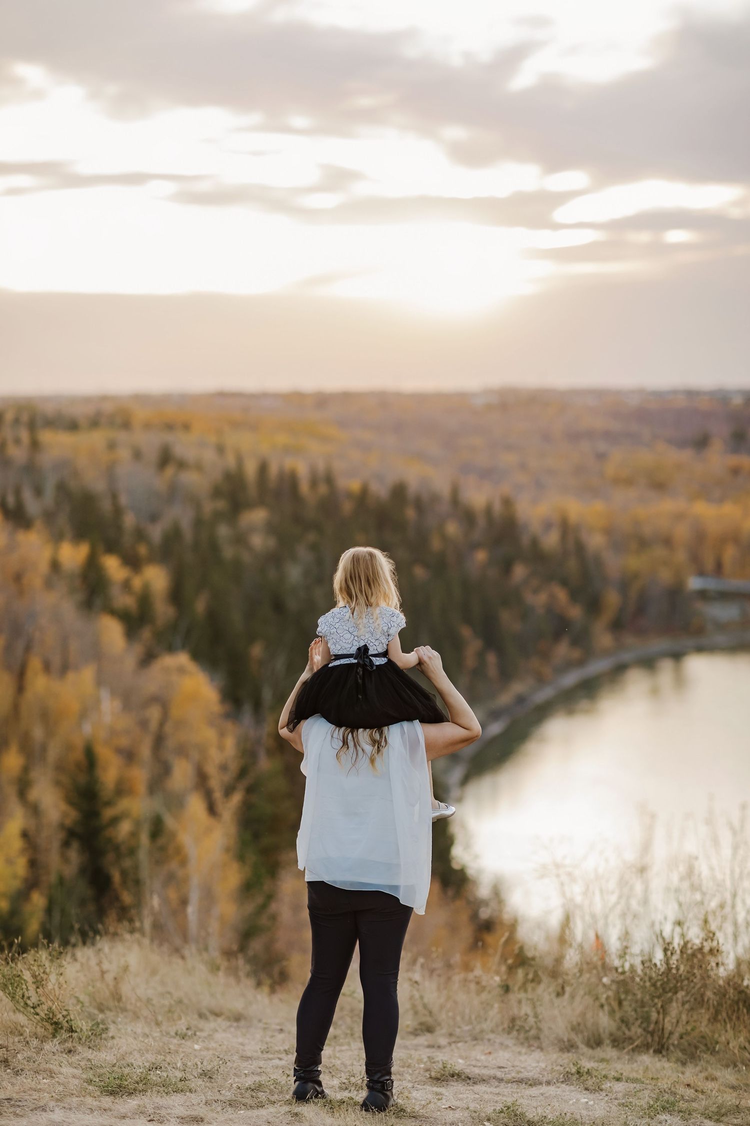 A mother carries her young daughter on her shoulders overlooking a river valley with autumn foliage at sunset.