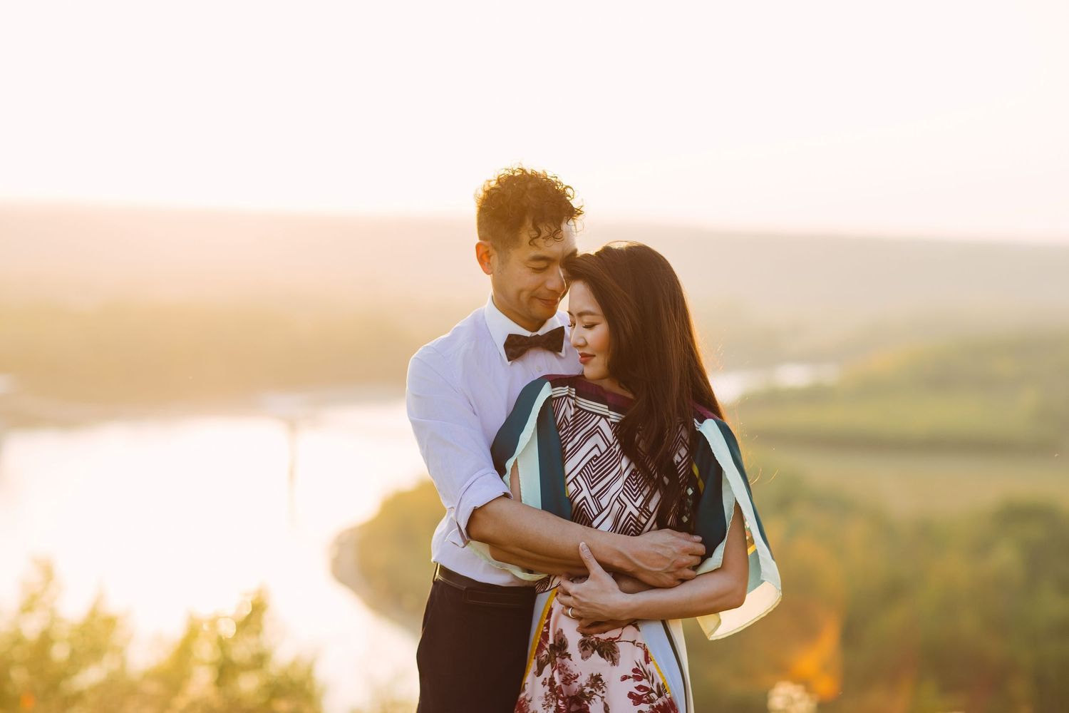 Couple embracing at golden hour on a hilltop overlook with a river and lush landscape in the background.