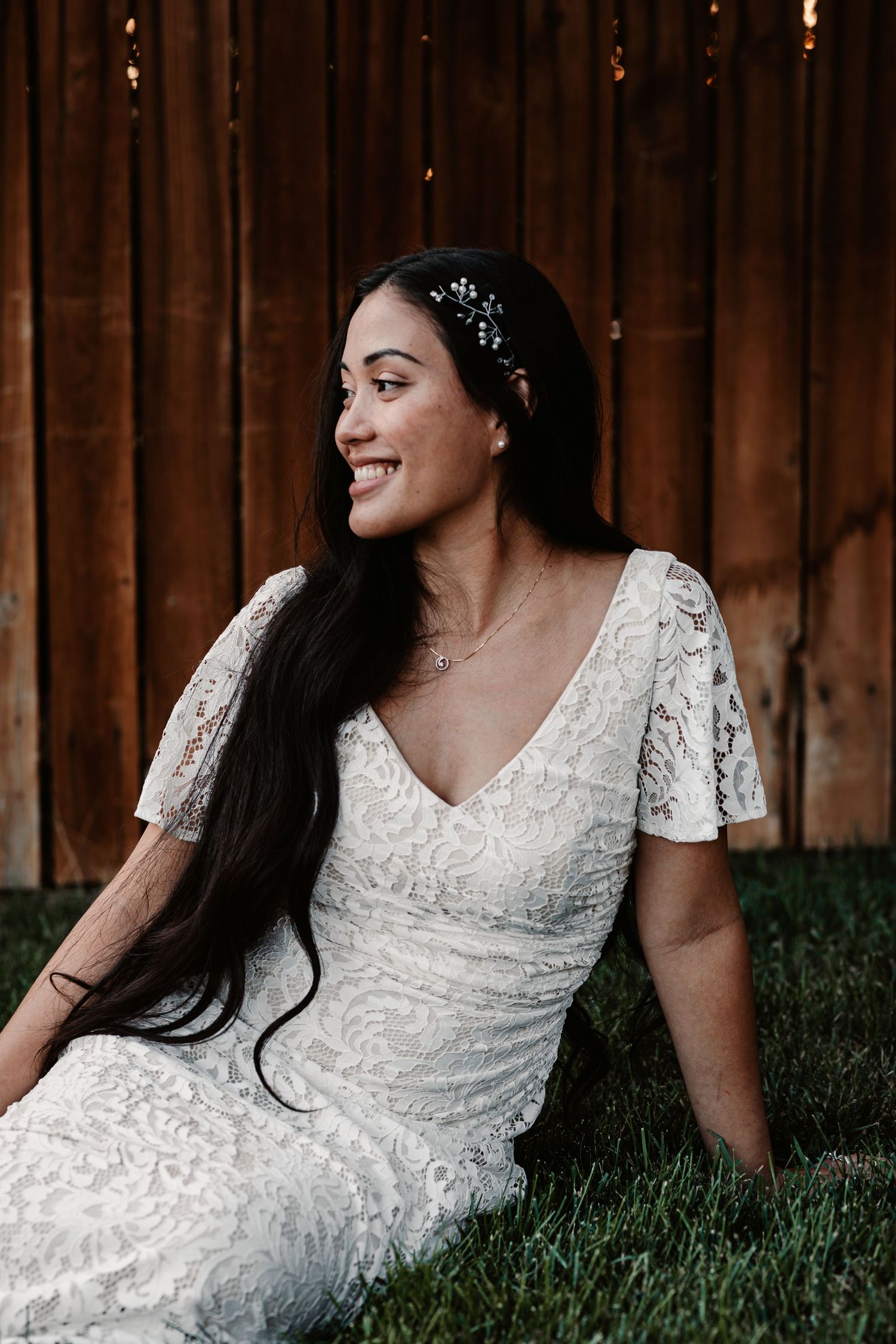 Smiling bride in white lace dress with flutter sleeves and floral hair accessory sitting on grass by wooden fence.