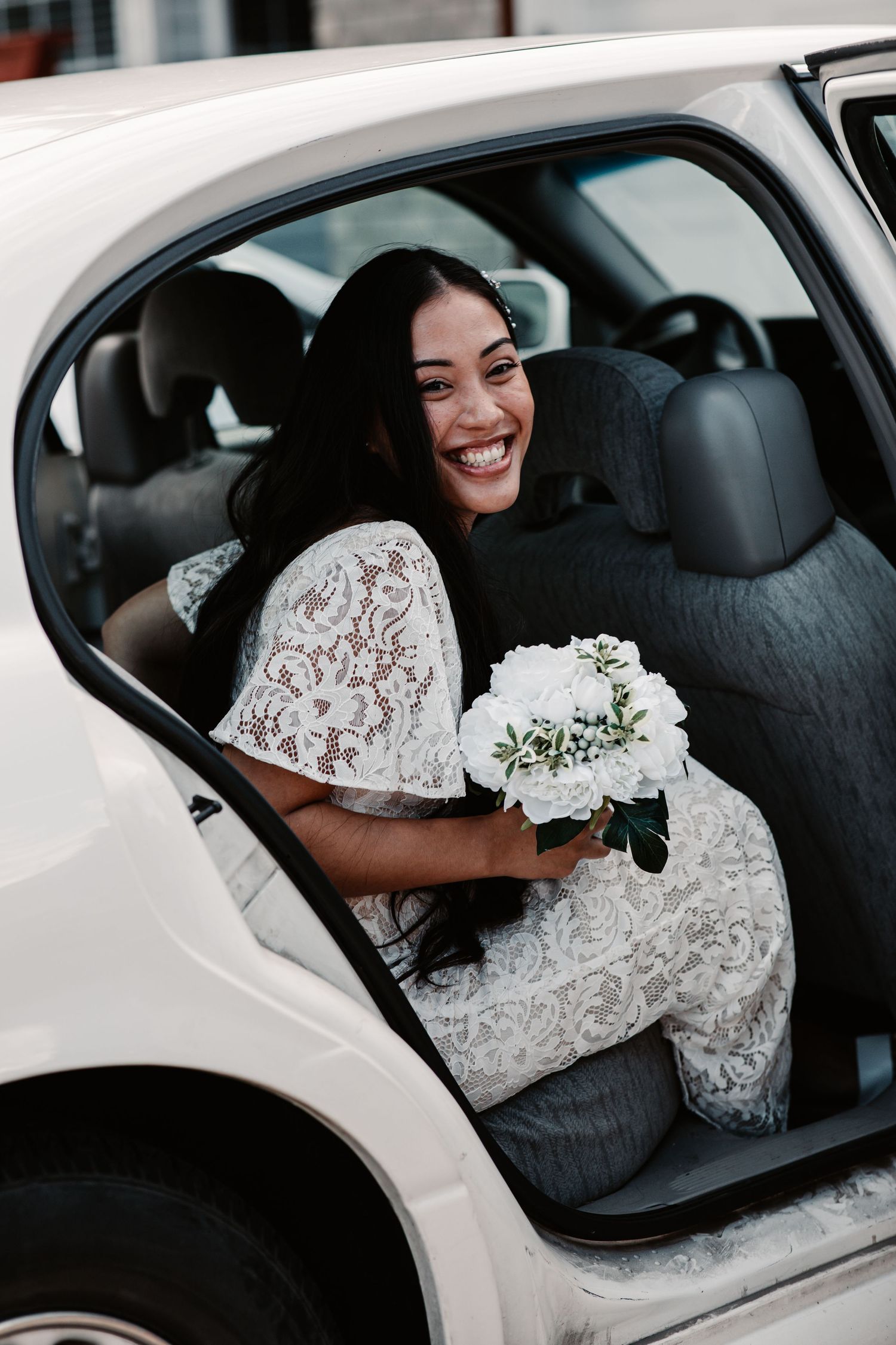 Smiling bride in lace dress holding white bouquet while sitting in a luxury white car on her wedding day.