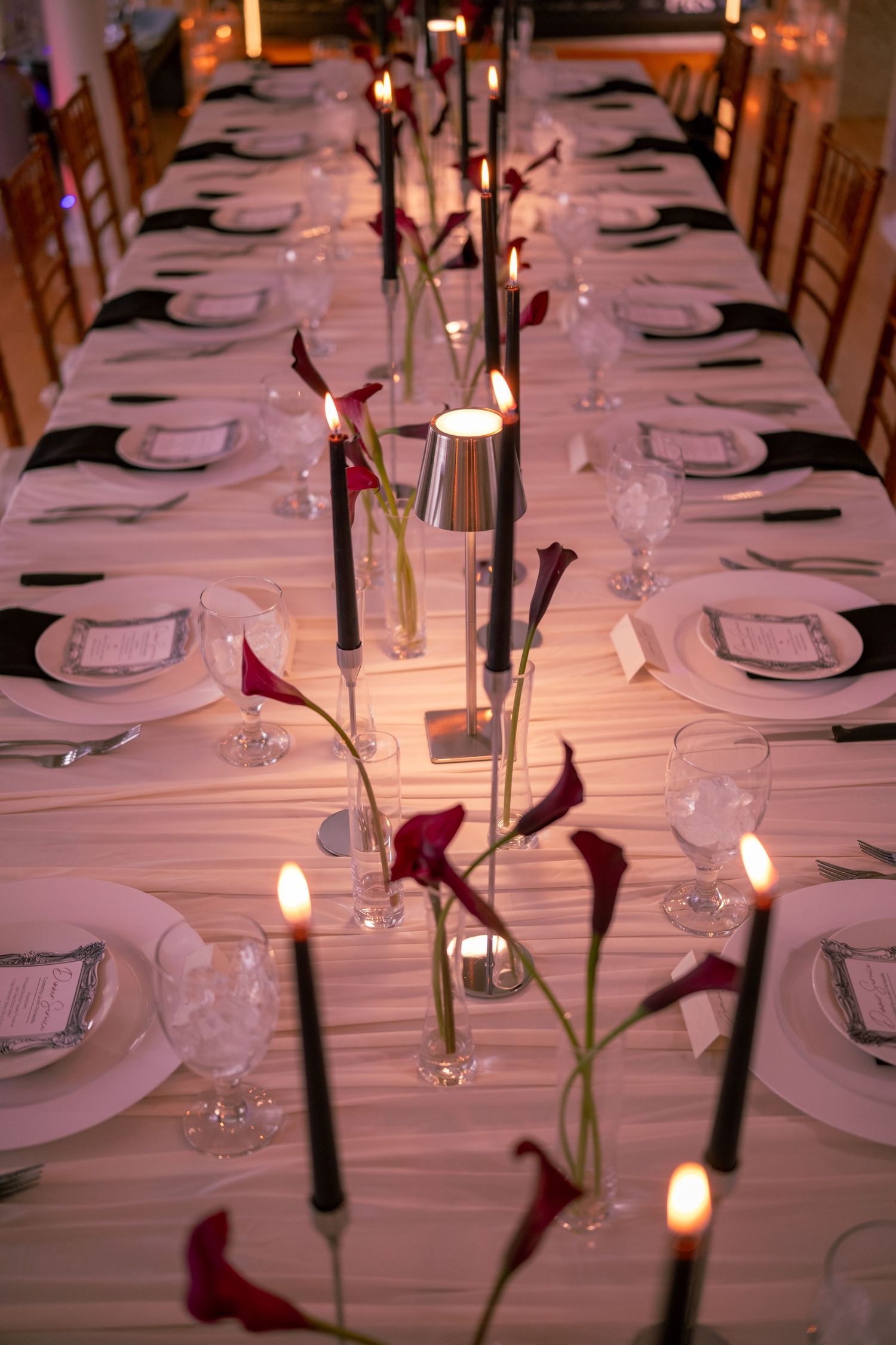 Elegant long banquet table set with dark calla lilies, black candles, and white place settings.