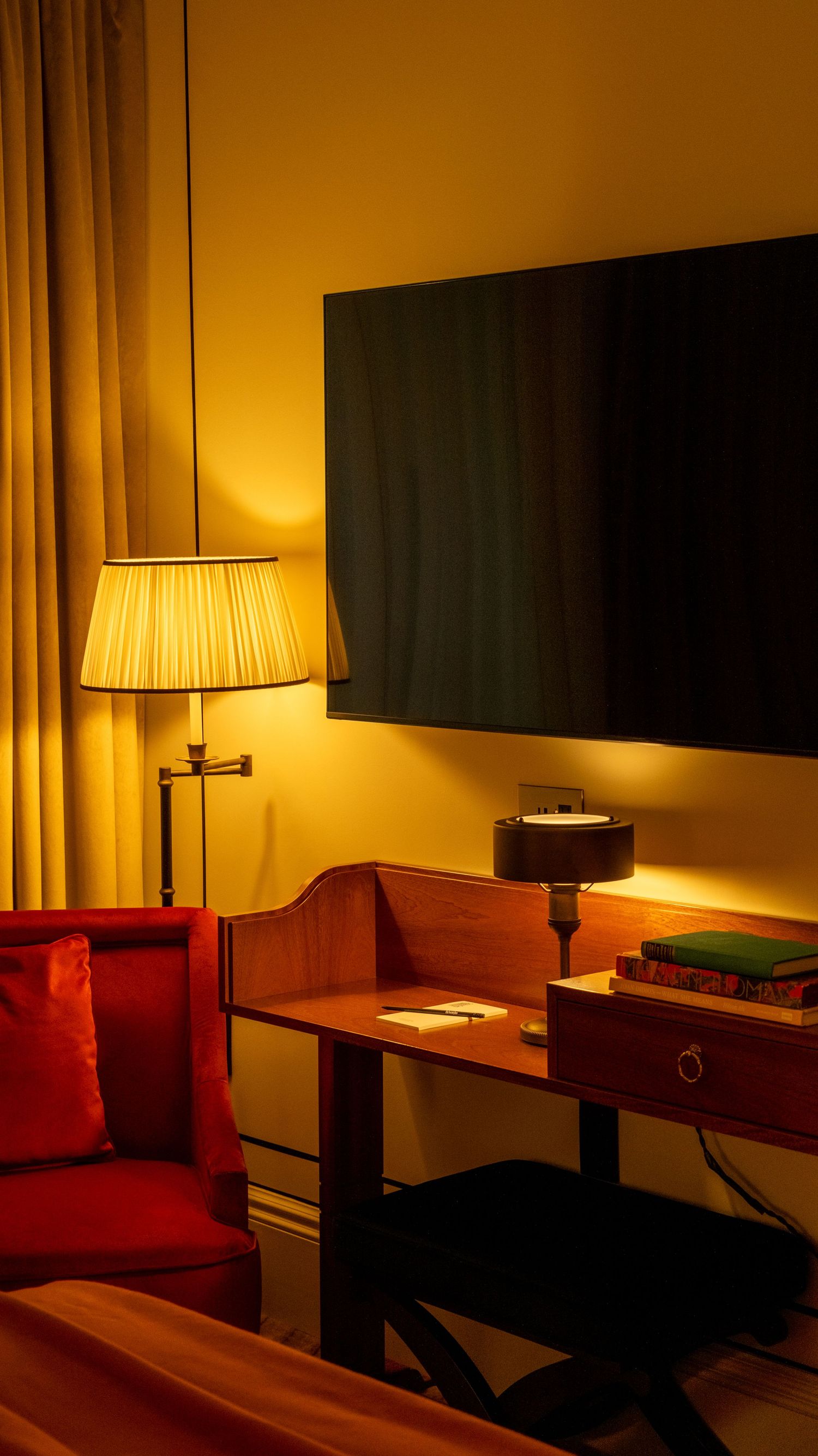 A warmly lit hotel room with a wooden desk, pleated floor lamp, red chair, and wall-mounted TV.