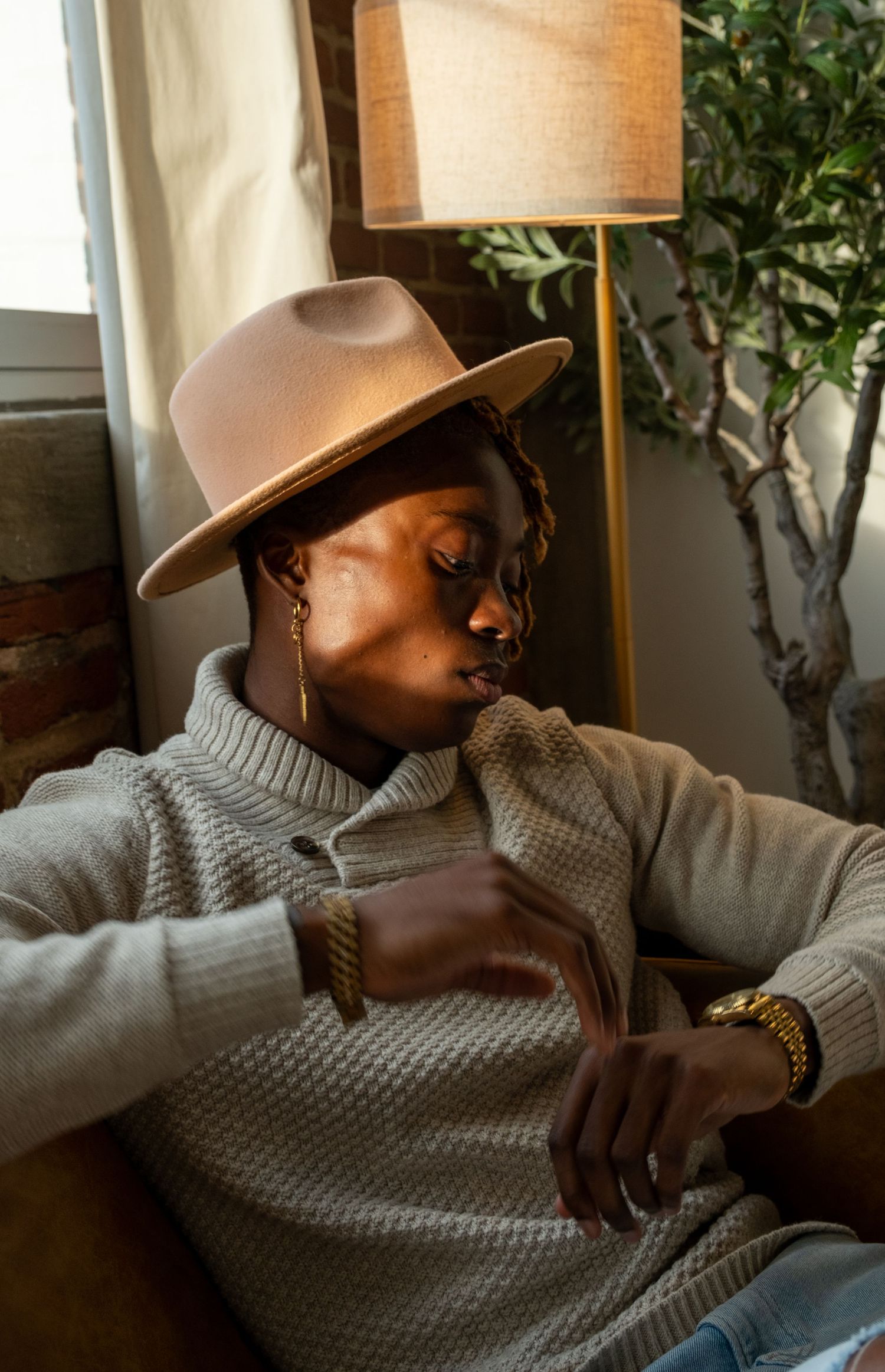 Person in cream knit sweater and tan hat relaxing on a couch near a lamp and indoor plant.