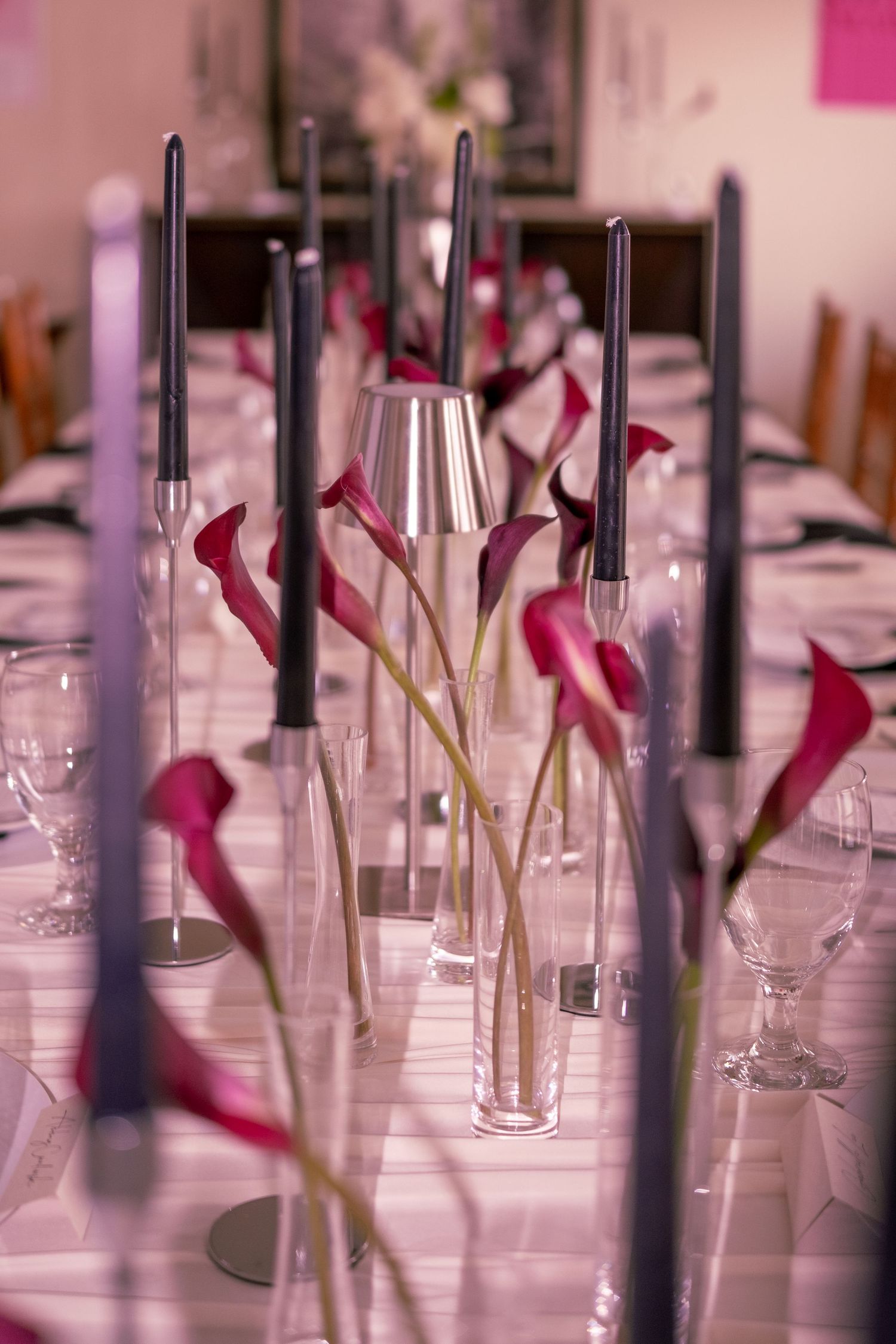 Elegant table setting with tall dark candles and pink calla lilies in glass vases for a formal dinner.