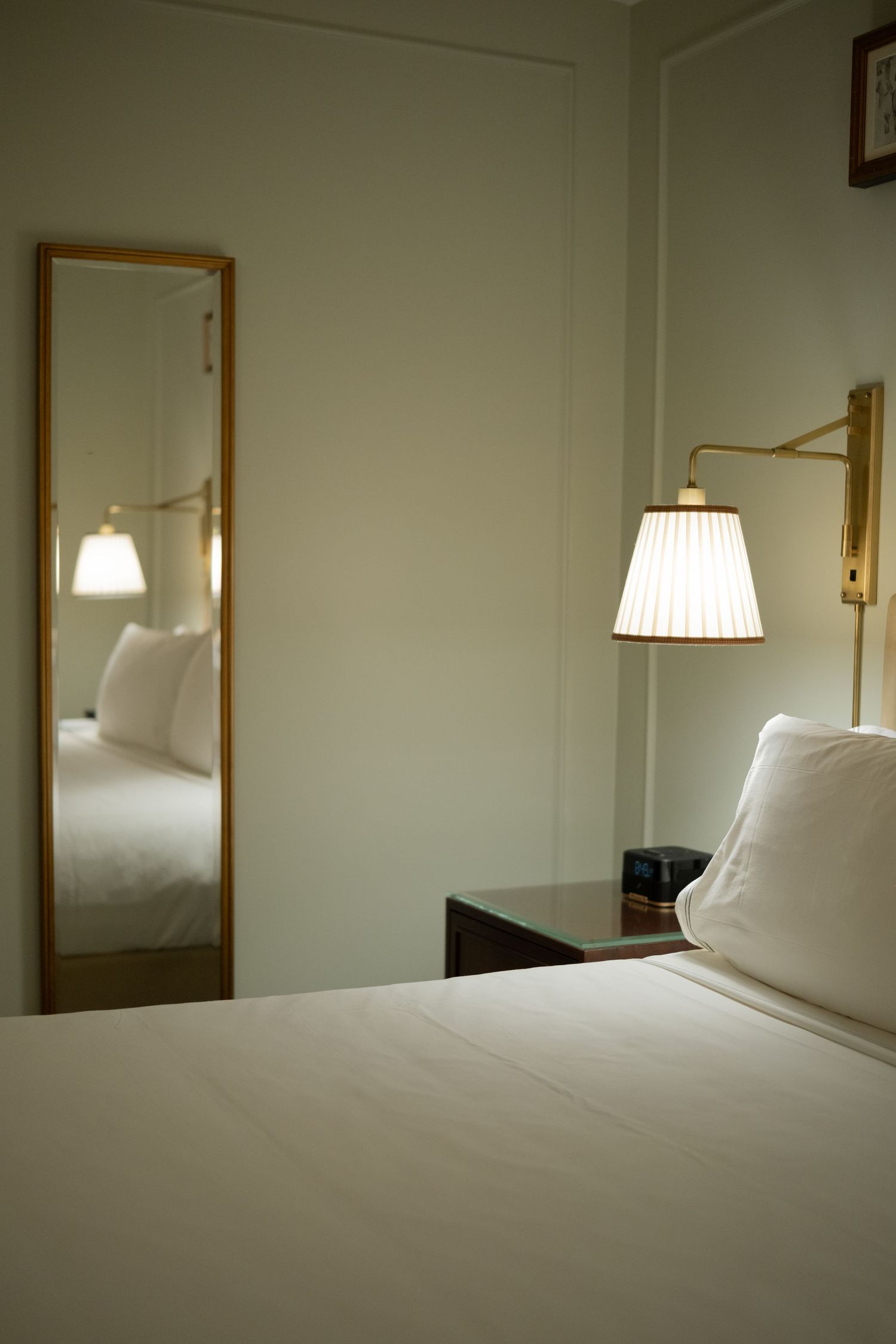 Minimalist hotel bedroom with white bedding, gold-framed mirror, and warm wall-mounted lamp.
