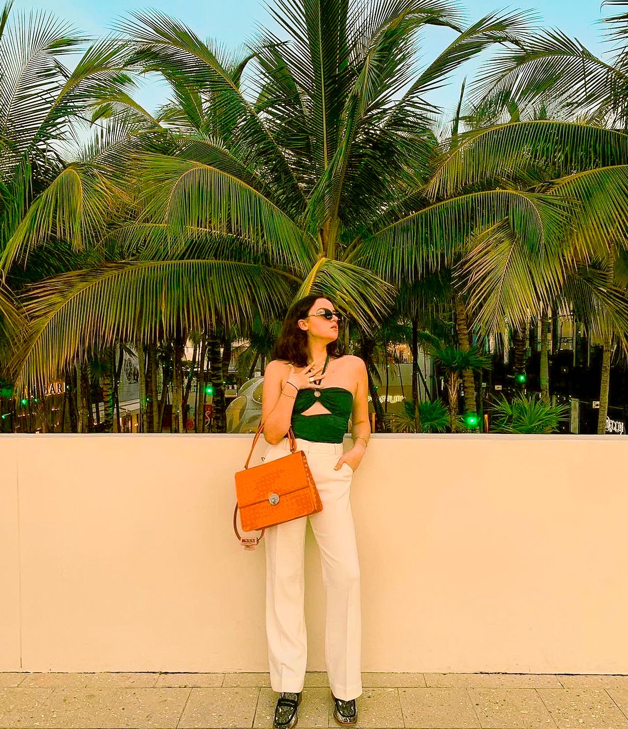 Woman in green crop top, white pants, and orange handbag posing before lush tropical palm trees.