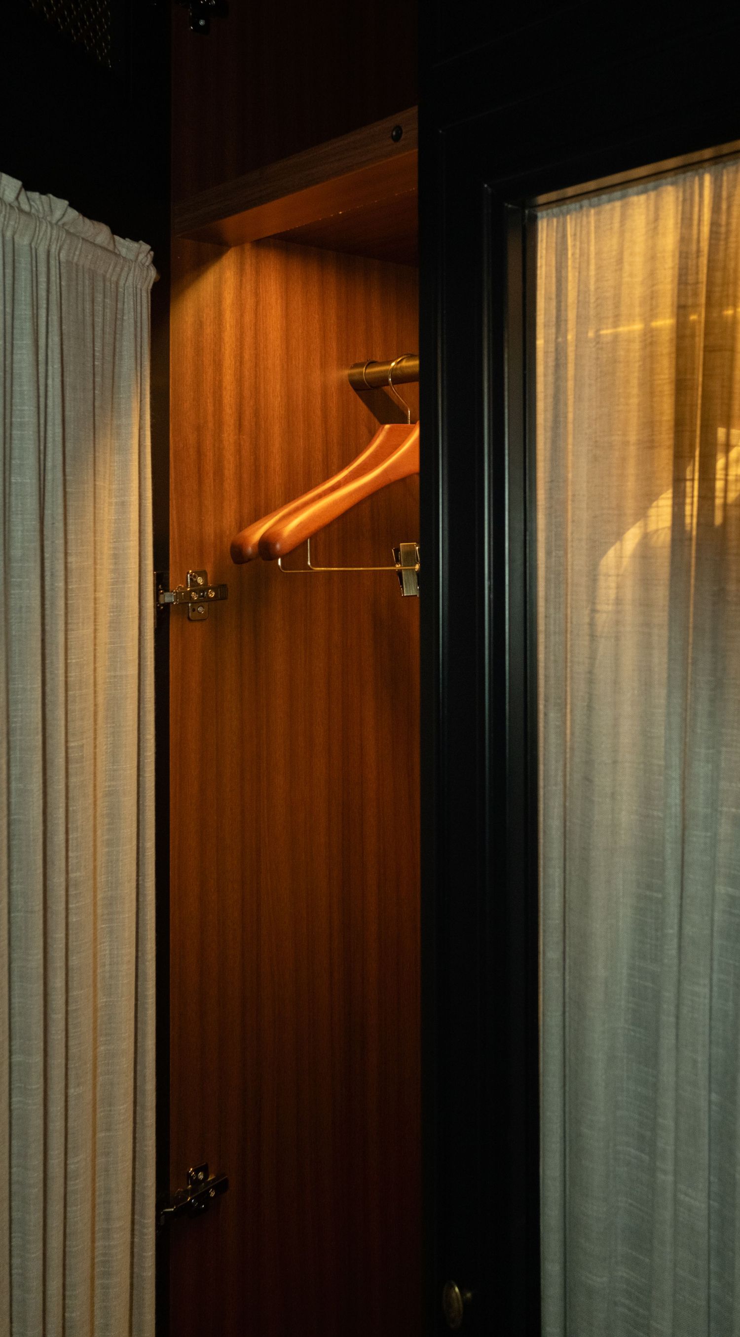 Warmly lit wooden wardrobe with vintage brass coat hangers between sheer linen curtains.