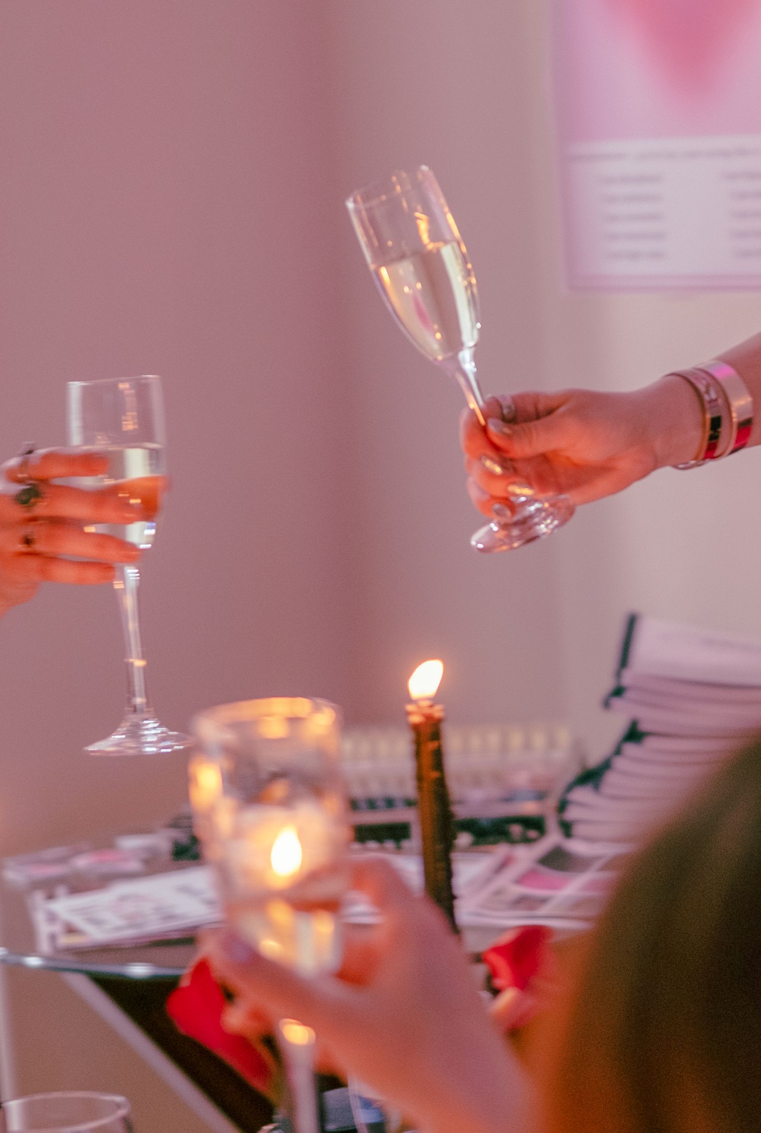 Two hands holding champagne flutes in a toast over a candlelit table at an elegant dinner party.