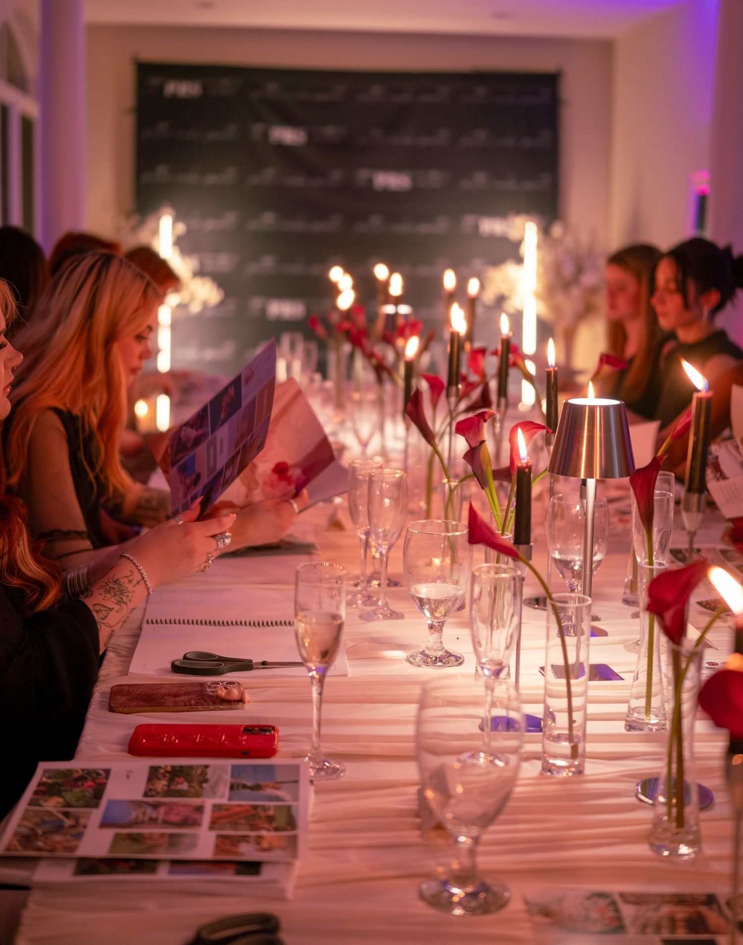 Elegant candlelit dinner party with guests reading menus along a long decorated table with red flowers.