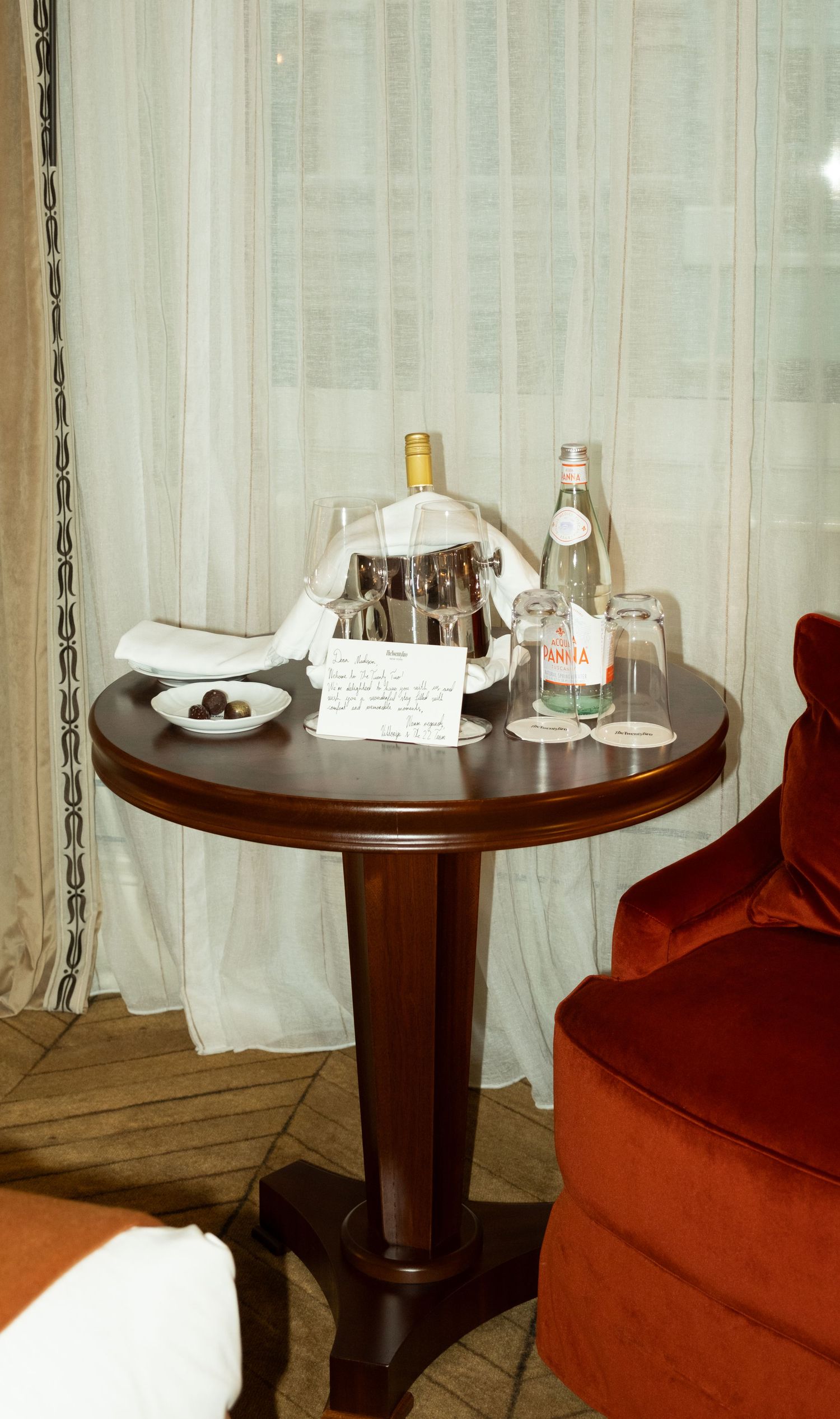 Elegant hotel room side table with wine bottle in ice bucket, water bottle, and welcome note by red velvet chair.