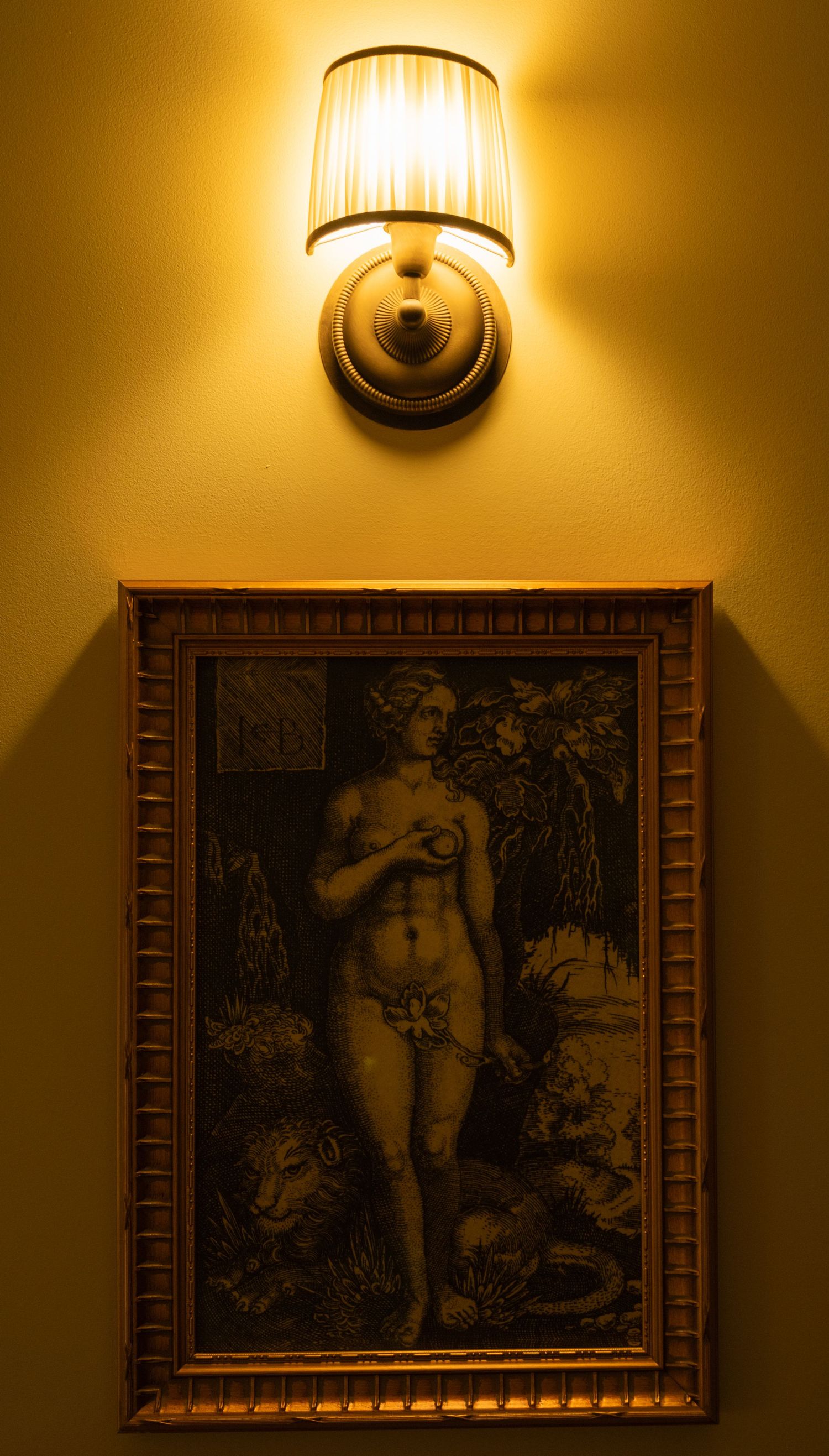 Elegant wall lamp casting warm light over a framed classical nude artwork on a yellow wall.