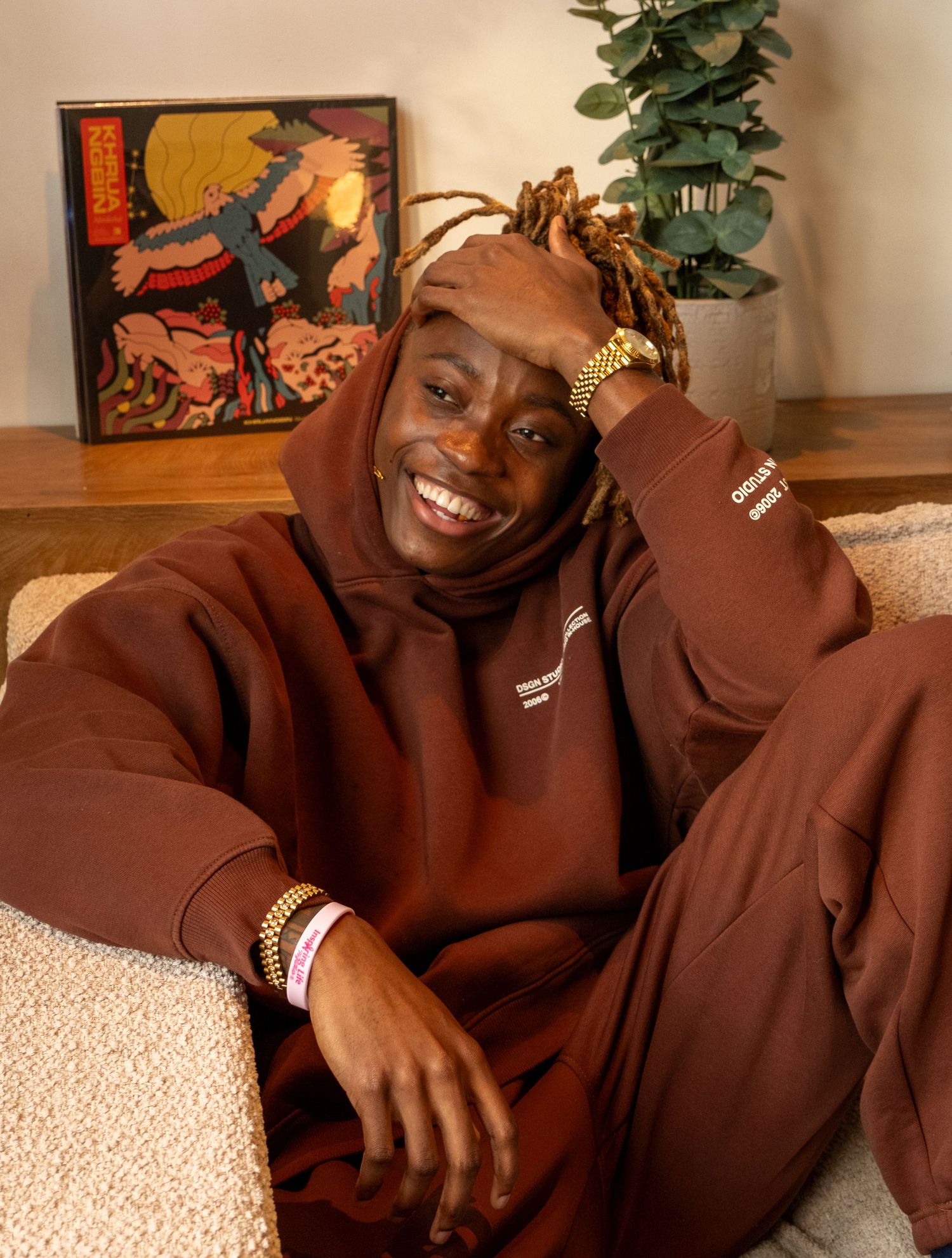 Person in brown hoodie and sweats smiling while relaxing on a couch with colorful art in background.