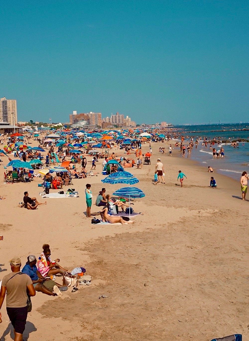 Crowded summer beach with colorful umbrellas, sunbathers on sandy shore, and city skyline under clear blue sky.