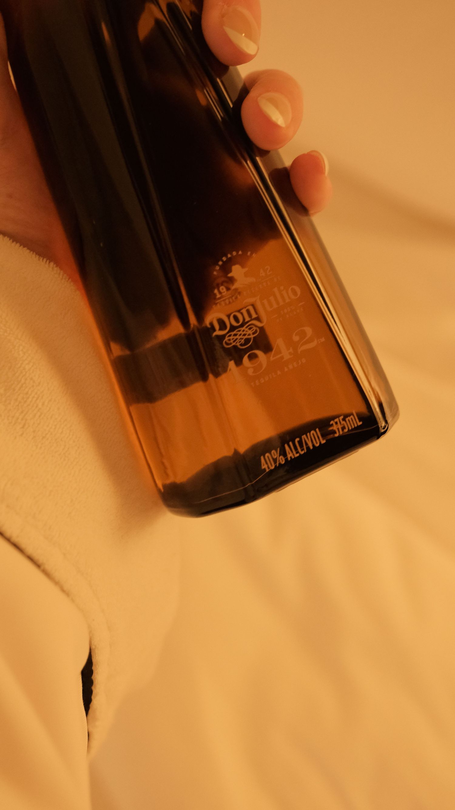 Hand holding a Don Julio 1942 tequila bottle on white hotel bedding.