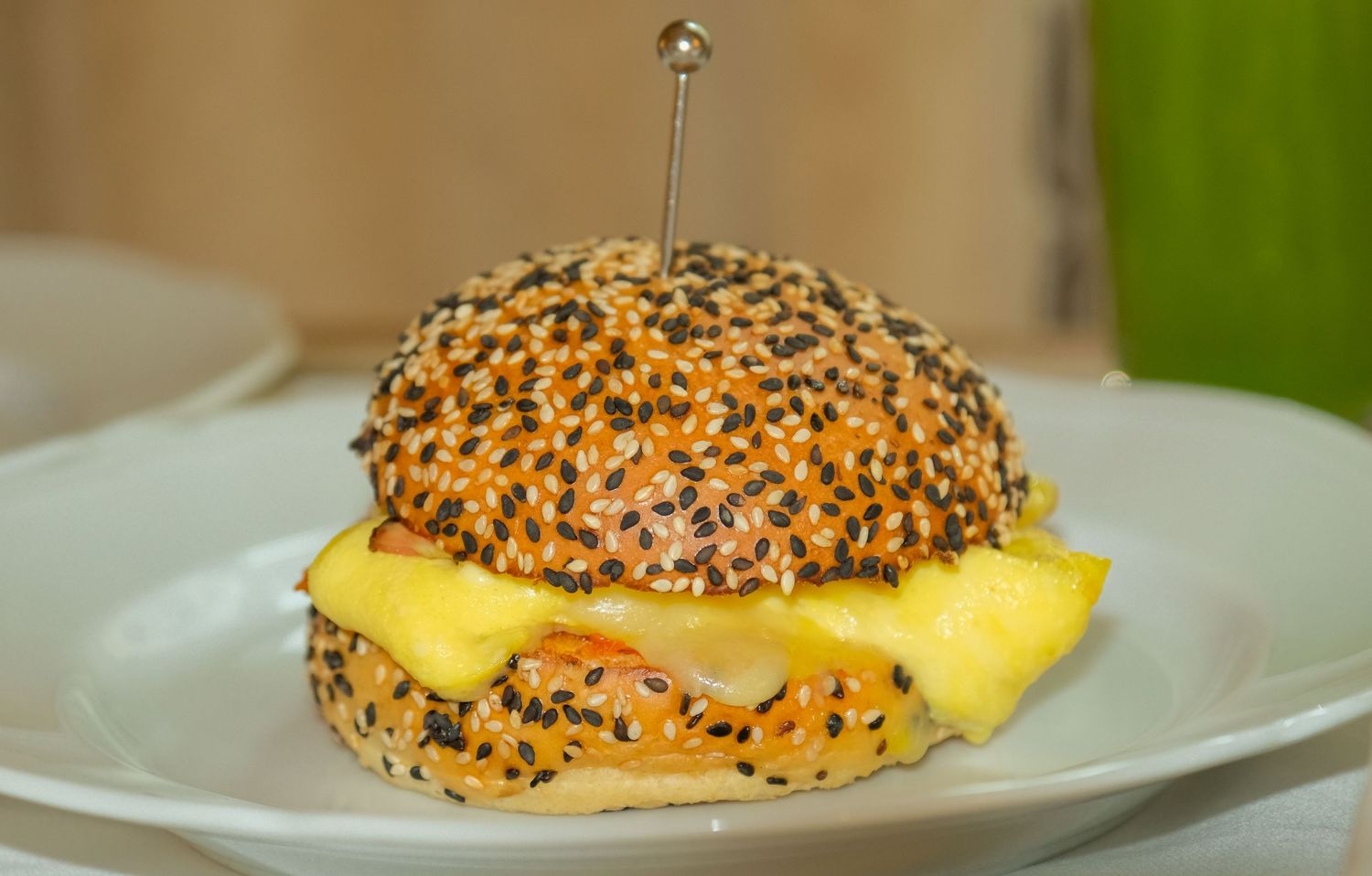 Gourmet sesame seed bun sandwich with melted cheese on white plate, skewered with cocktail pick.