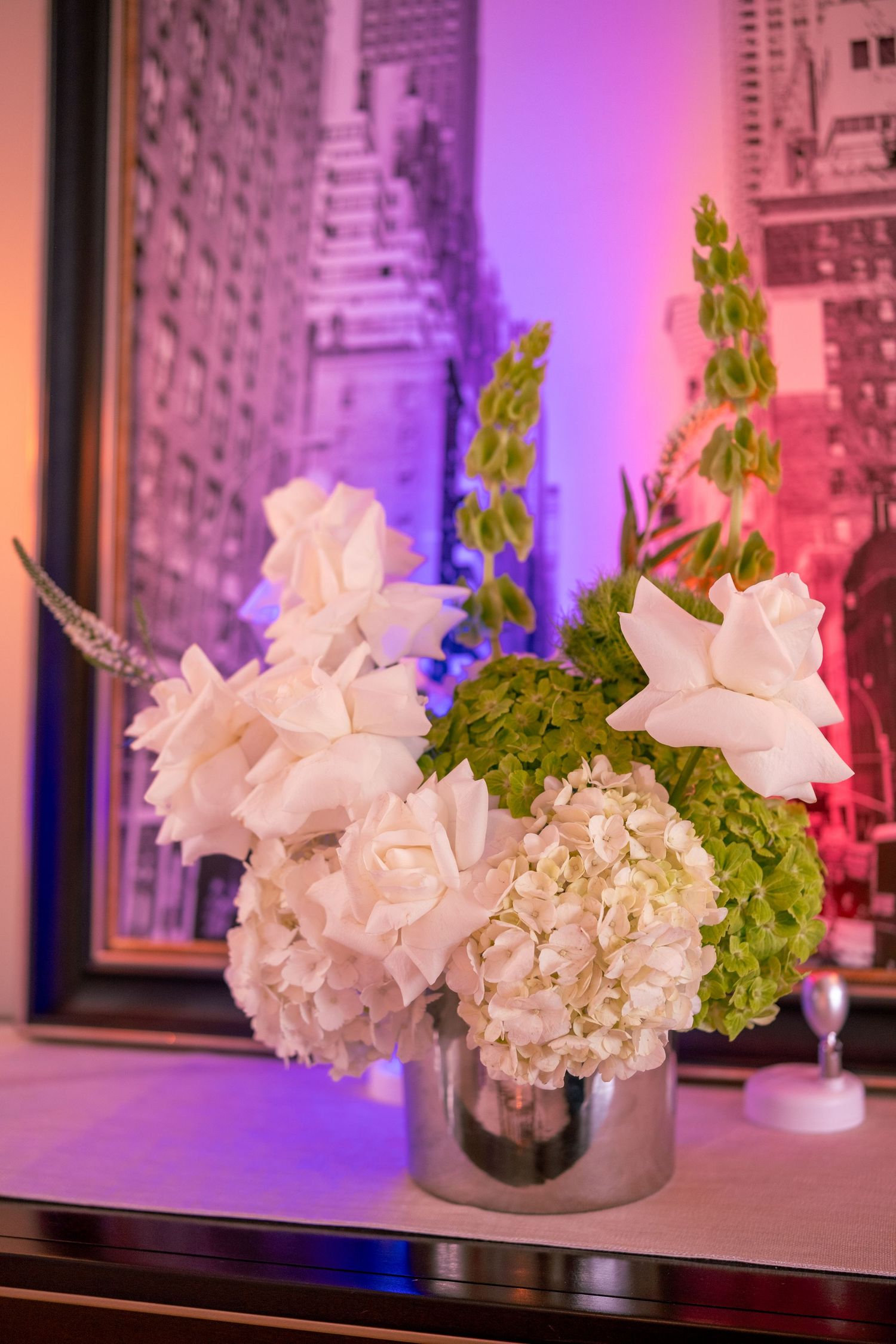 White floral arrangement with hydrangeas and roses in a silver vase before a framed city artwork.