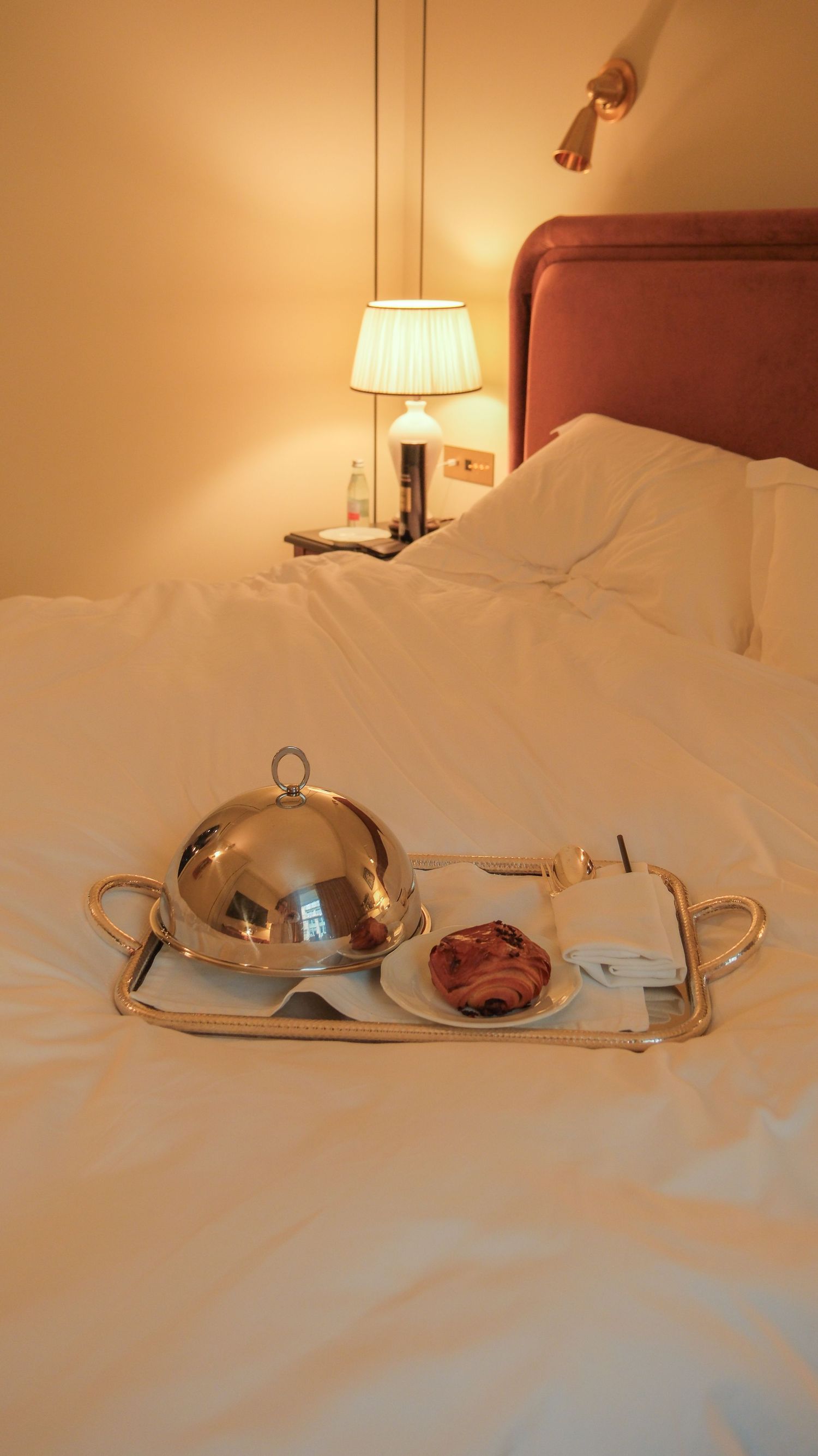 Silver domed room service tray with pastry on white bed in warmly lit luxury hotel room.