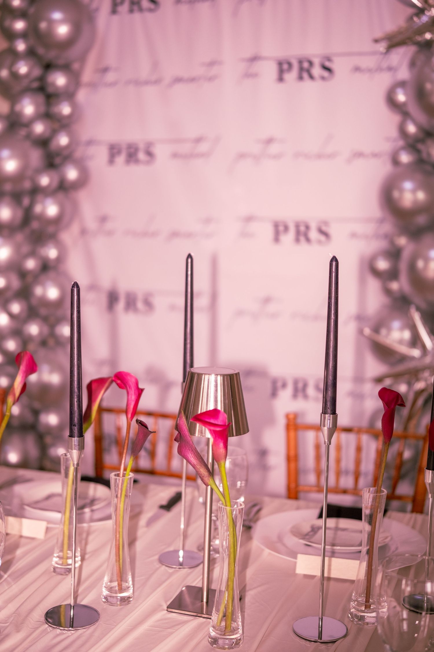 PRS branded dinner table with dark taper candles, pink calla lilies, and silver balloon arch backdrop.