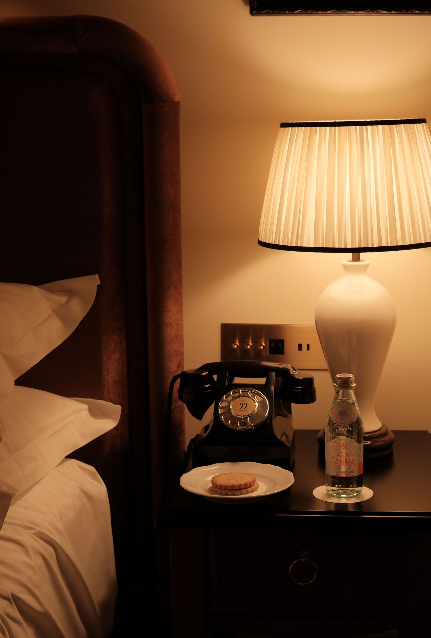 Vintage black rotary phone on dark wood nightstand beside glowing lamp and water bottle in cozy hotel room.