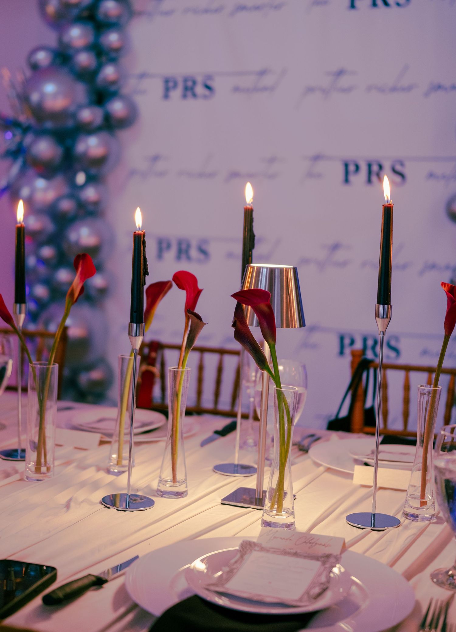 Elegant PRS event table with tall dark candles, red calla lilies, and branded step-and-repeat backdrop.