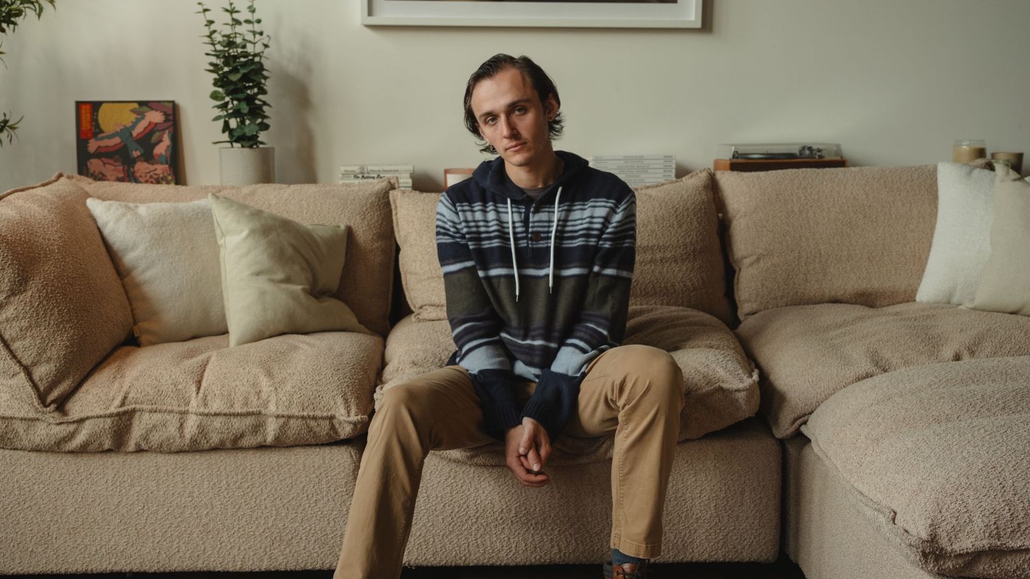 Slim young man in striped hoodie sitting alone on a beige couch in a cozy living room.