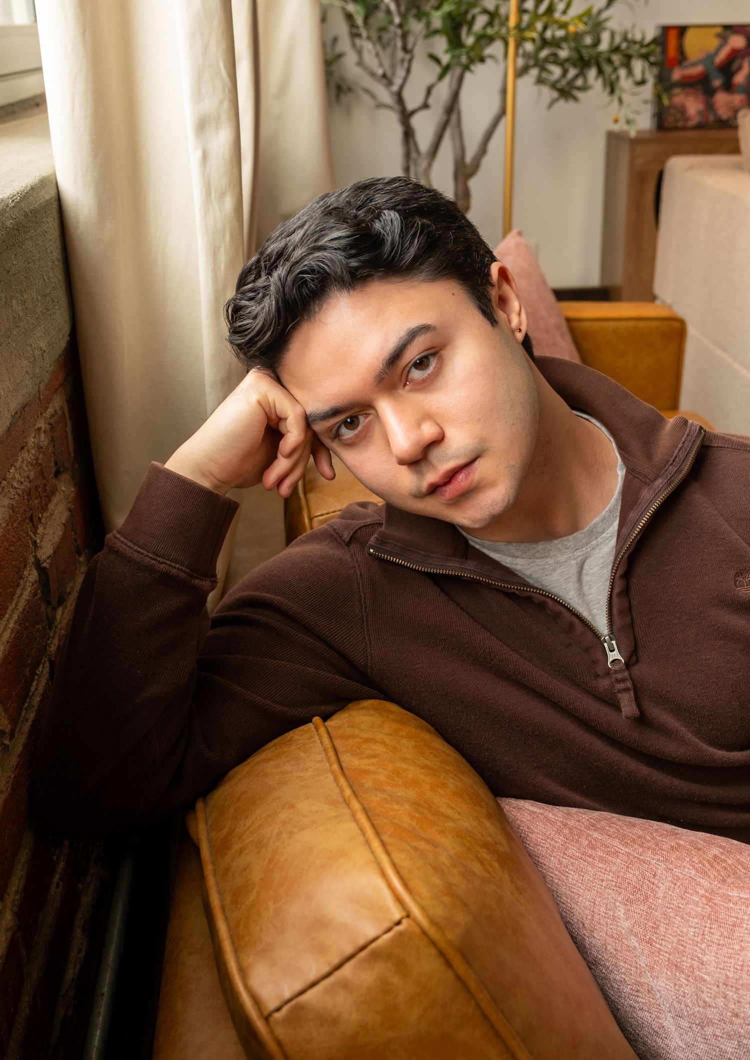 Young man in brown zip-up sweatshirt reclining on a leather couch with a pensive expression.