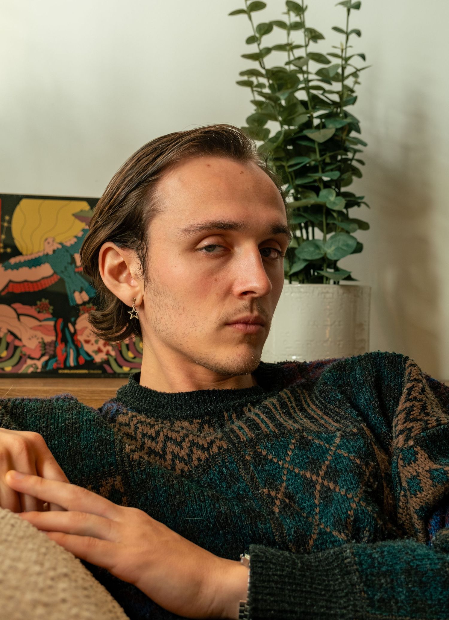 Young man in teal patterned knit sweater sitting on couch with colorful artwork and plant behind him.