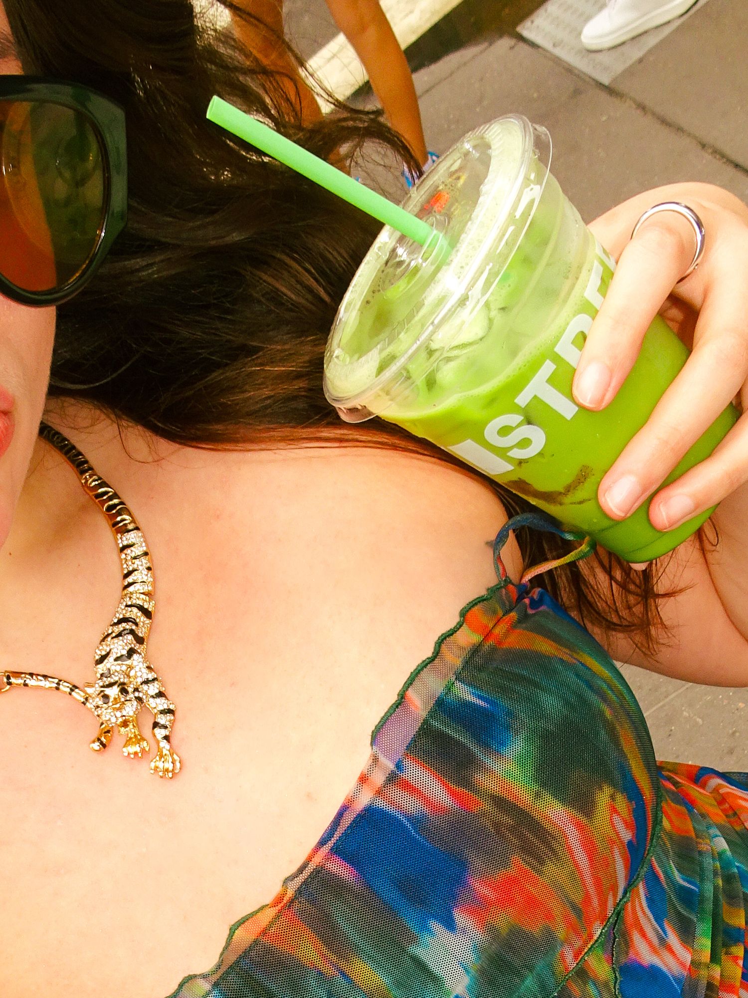 Woman holding iced matcha green tea drink with green straw, wearing tiger necklace and colorful top, sunglasses on head.