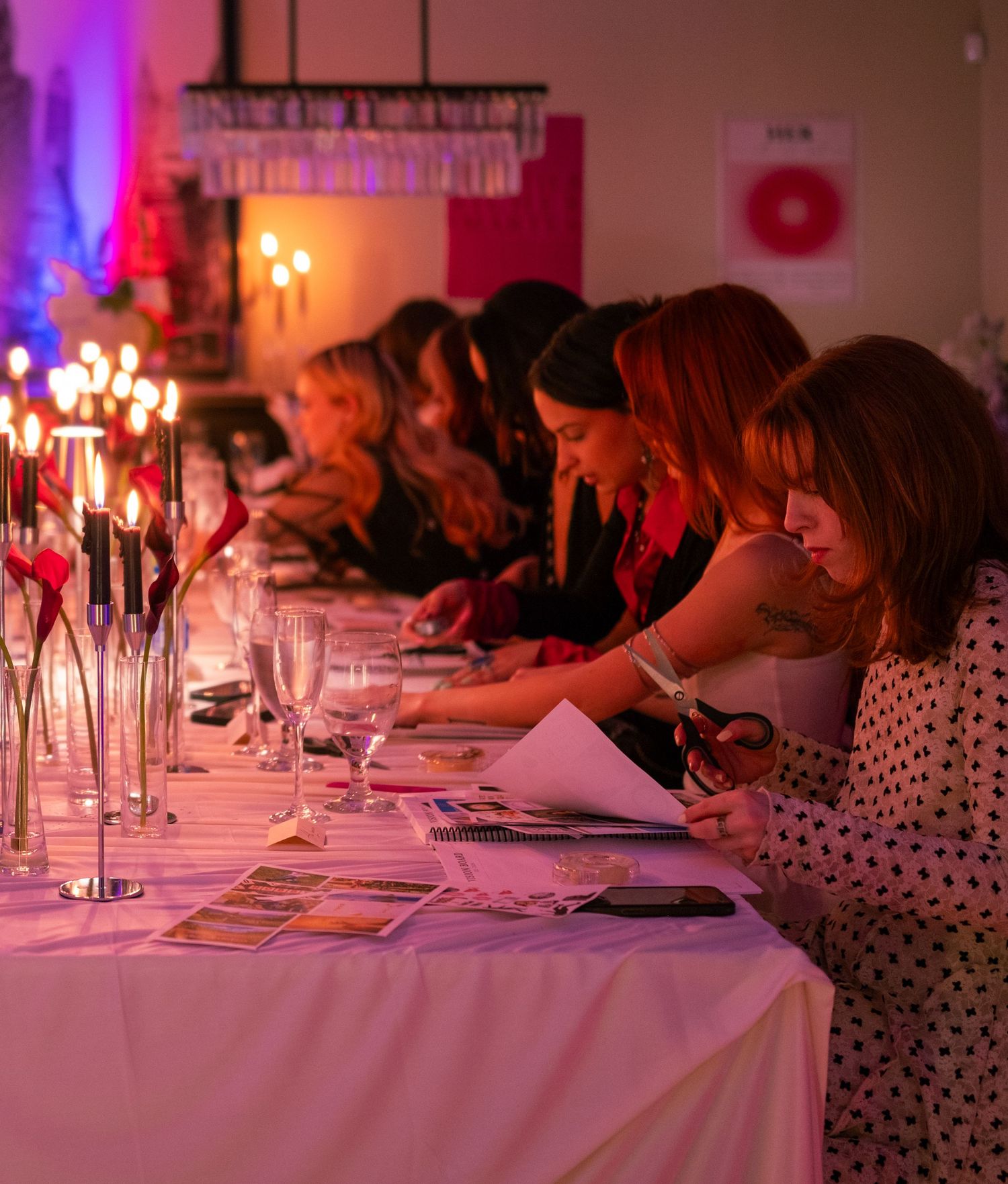 Row of women writing and reviewing papers at a candlelit event dinner table with glassware.