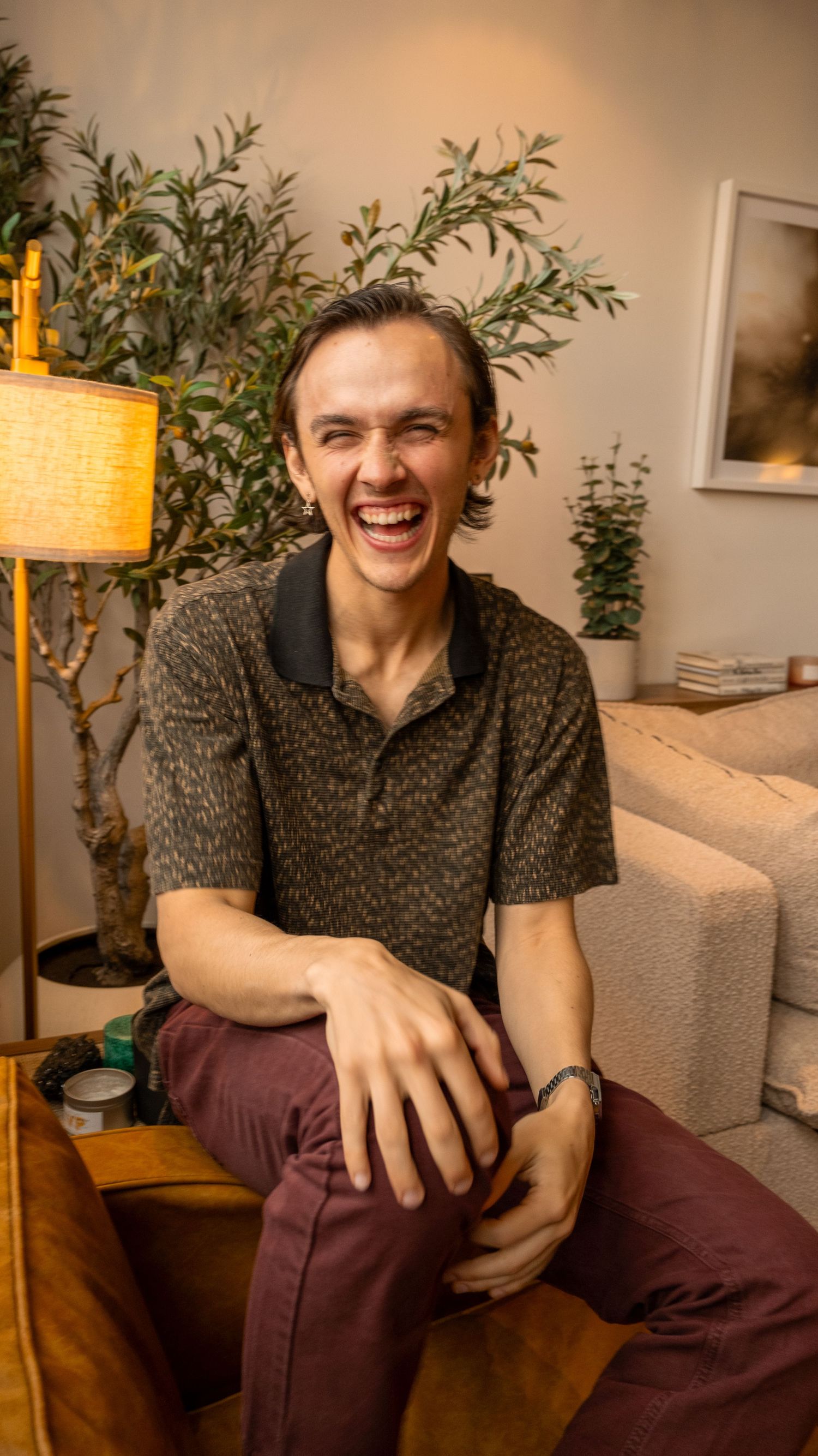 Young man laughing joyfully while seated on a wooden table in a cozy living room with plants.