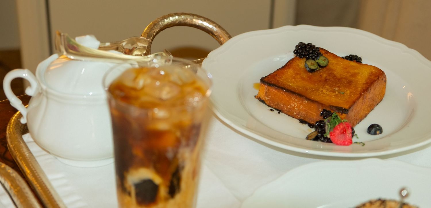 Golden French toast with blackberries on white plate beside iced coffee on elegant dining tray.