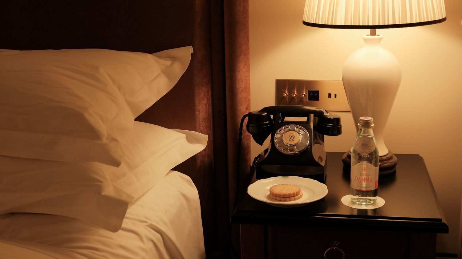 Cozy hotel bedside table with vintage black rotary phone, lamp, water, and turndown cookie.