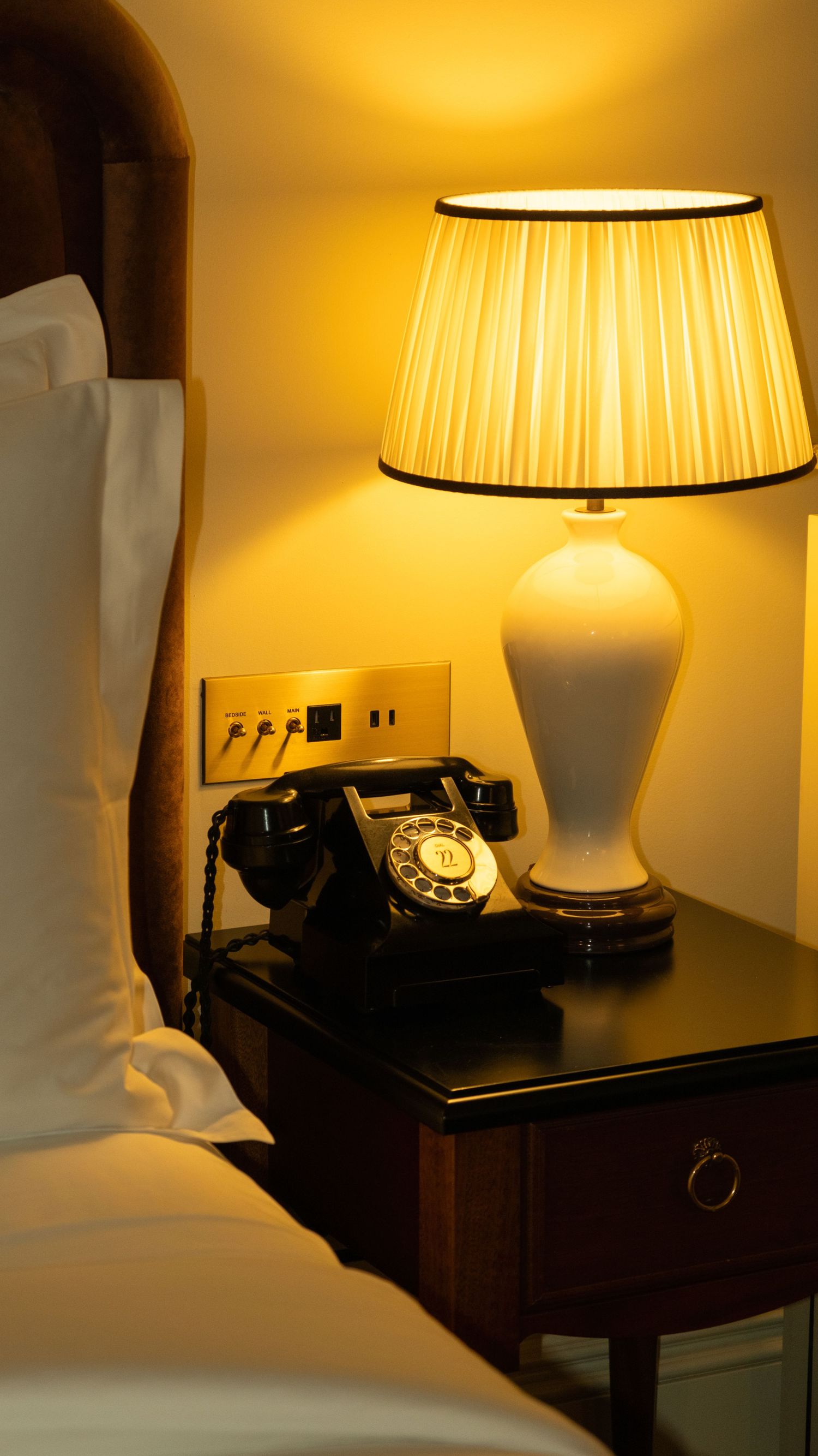 Vintage rotary telephone beside a glowing white ceramic bedside lamp in a luxury hotel room.