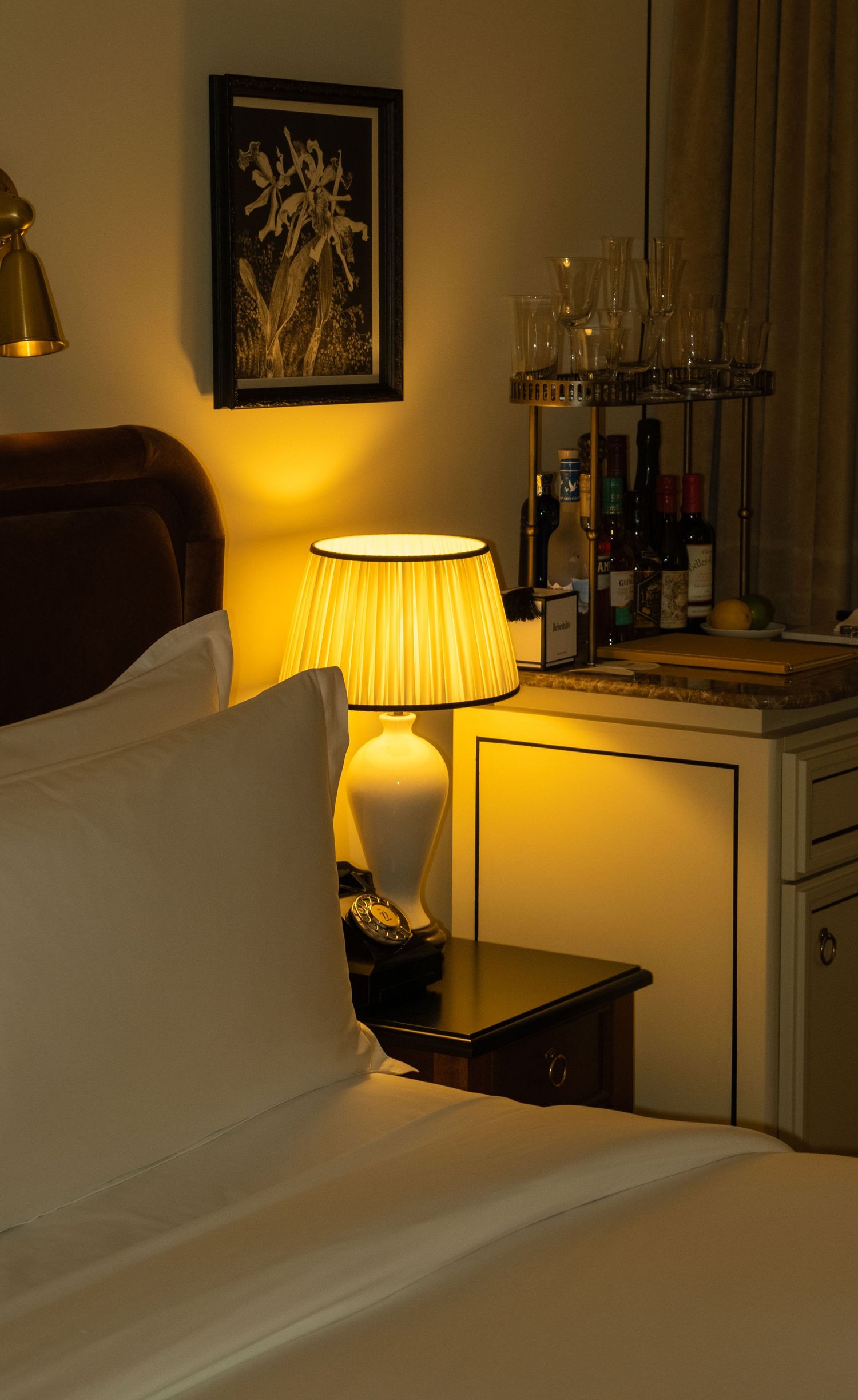 Cozy hotel bedroom with warm bedside lamp, rotary phone, and a stocked minibar in background.