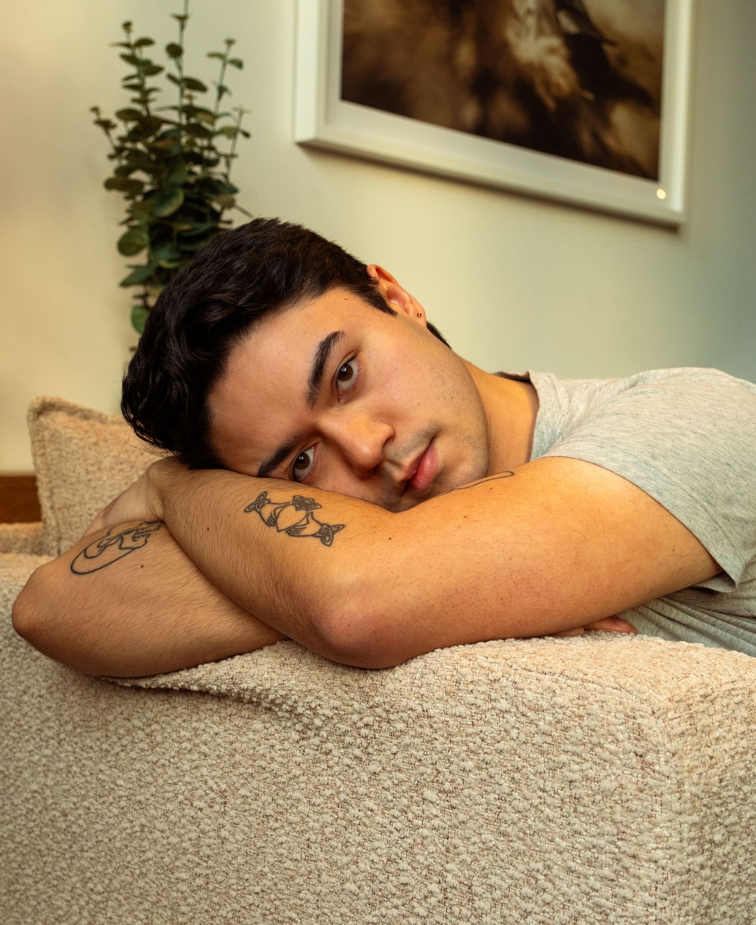 Young man with tattoos resting chin on arms over a beige sofa cushion.