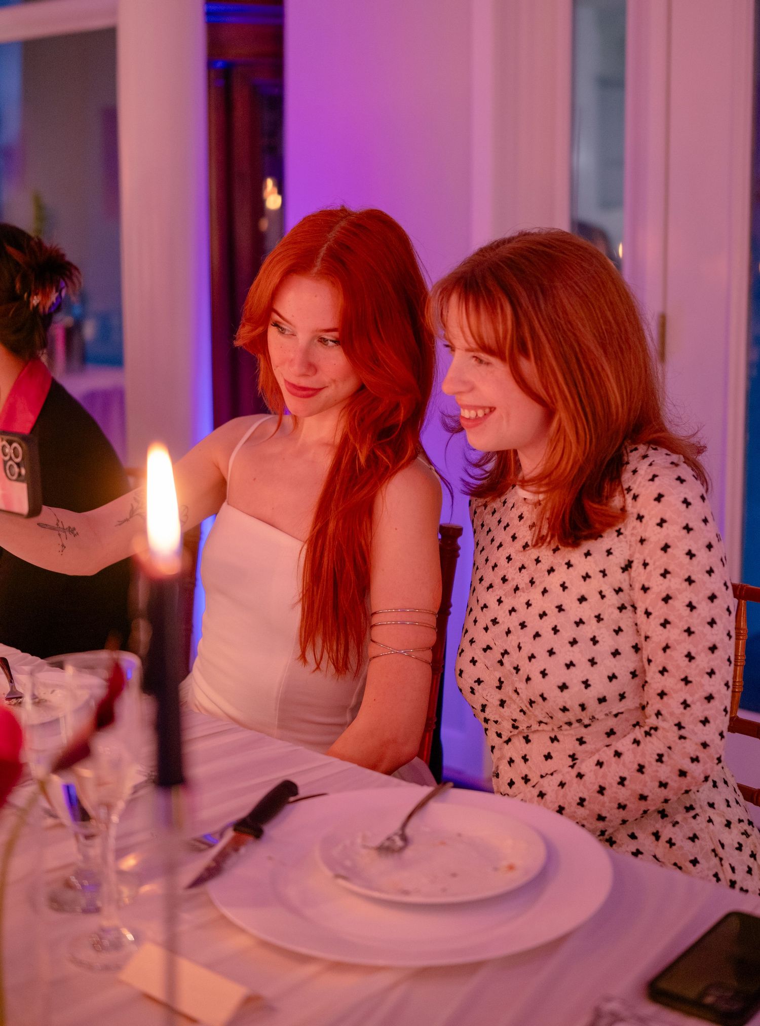 Two smiling women, one with red hair in white dress, seated together at a candlelit dinner event.