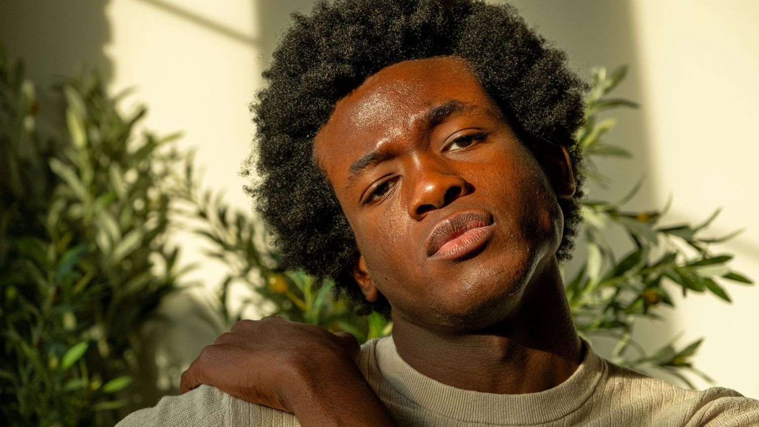 A young Black man with an afro in a beige top poses with sunlight and green plants in the background.