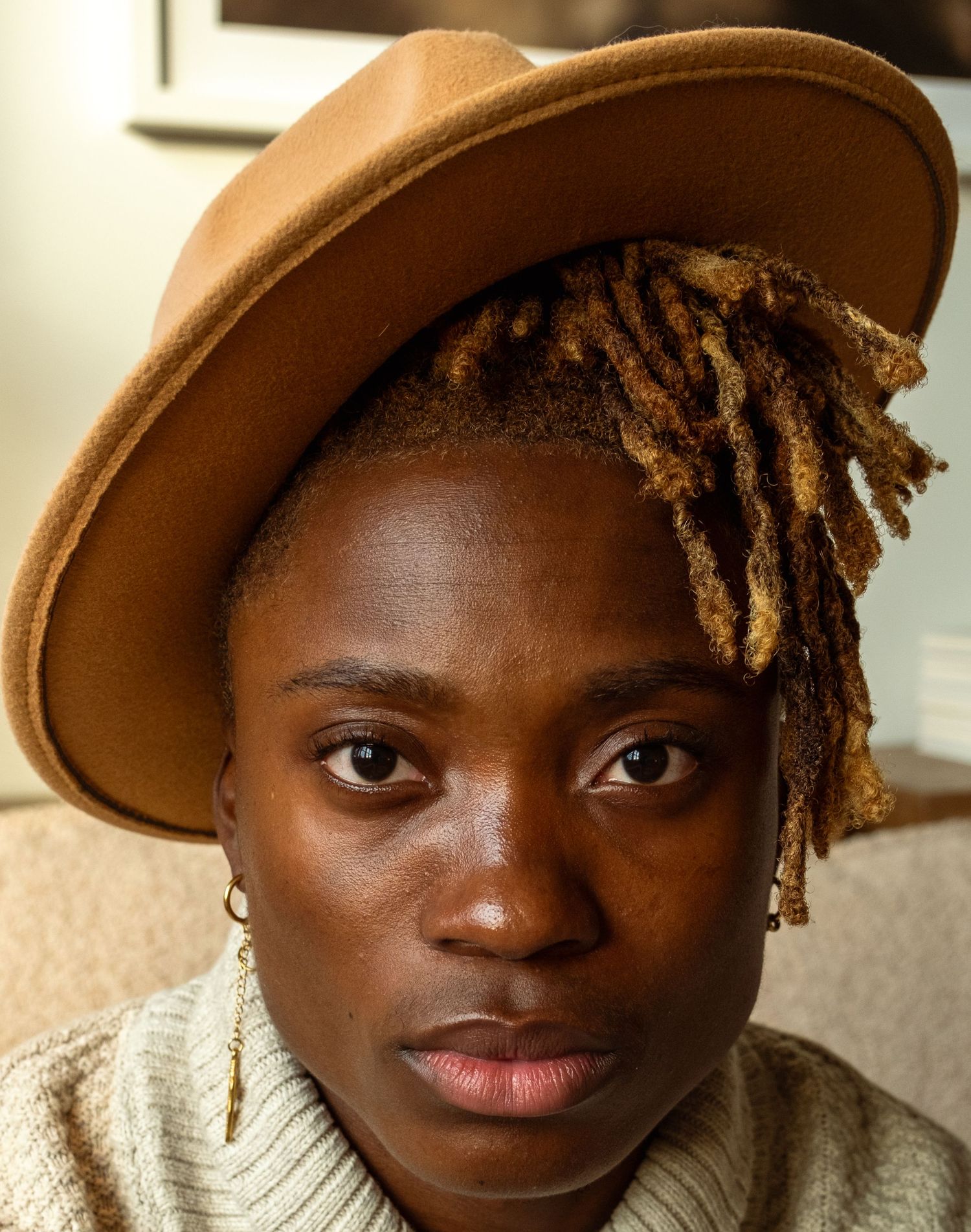 Close-up portrait of person with locs wearing a tan felt wide-brim hat and cream knit sweater.