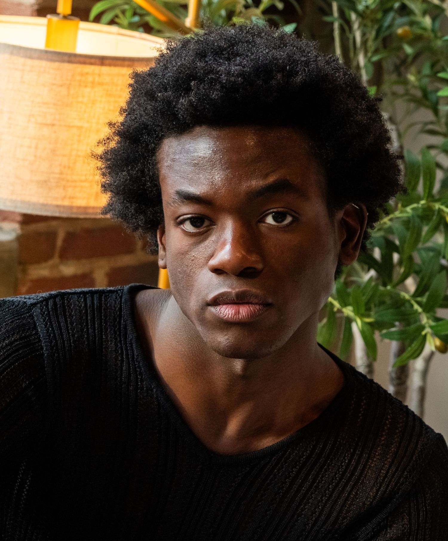 Young Black man in black sweater with natural afro hair posing near plants and warm lamp light.