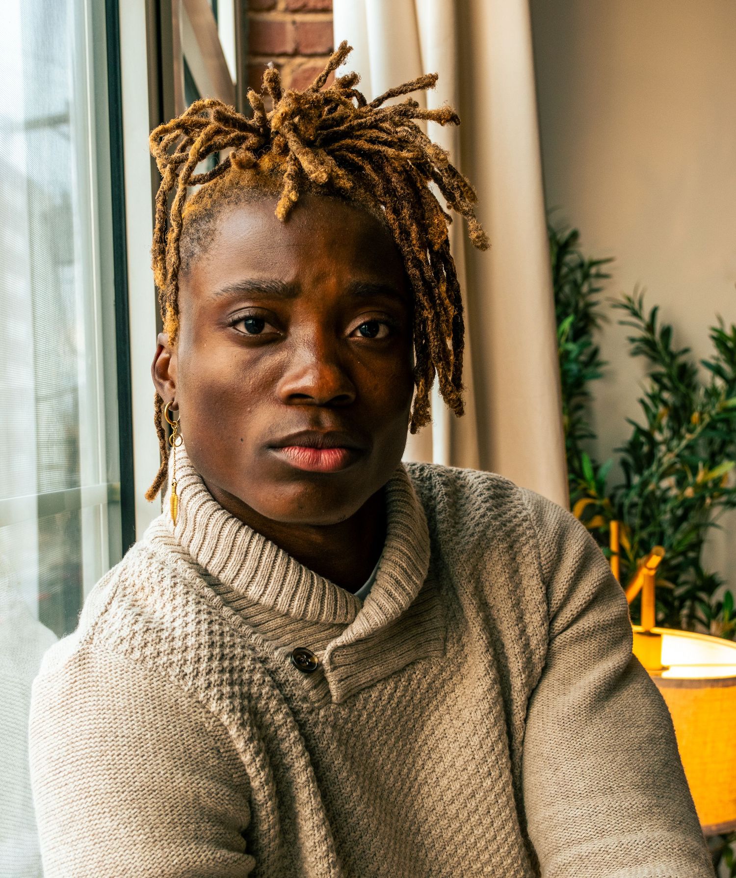A young Black man with blonde dreadlocks in a cream textured turtleneck sweater poses near a bright window.