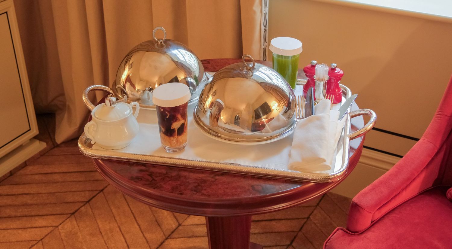 Elegant room service tray with silver domed covers, coffee cup, and condiments on ornate red side table.