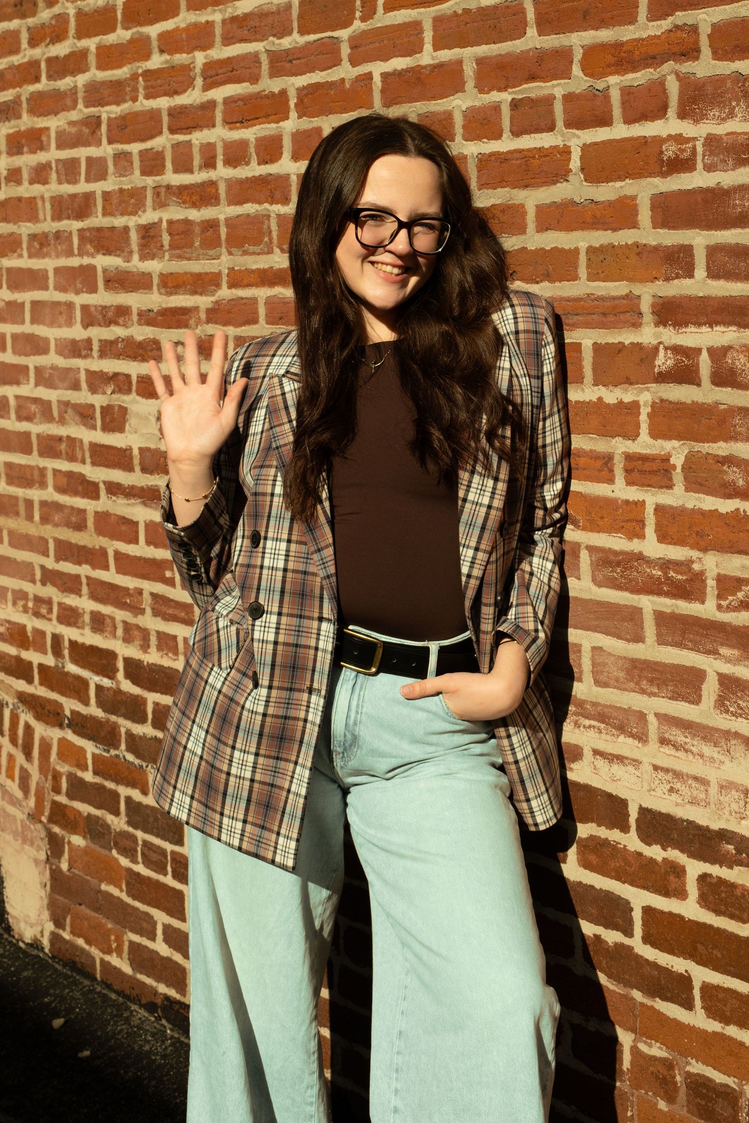 Woman in plaid blazer, brown top, and light jeans waves at camera against a sunny brick wall.