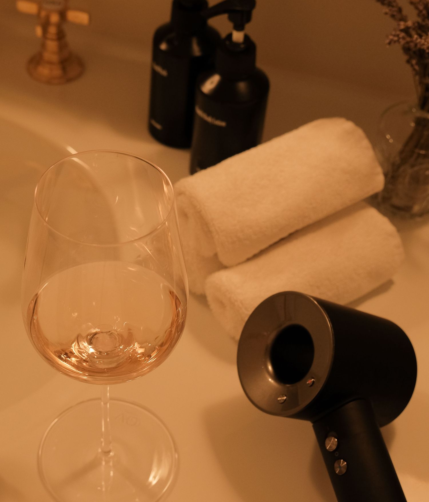 Rose wine glass beside a black hair dryer and rolled white towels on a hotel bathroom counter.
